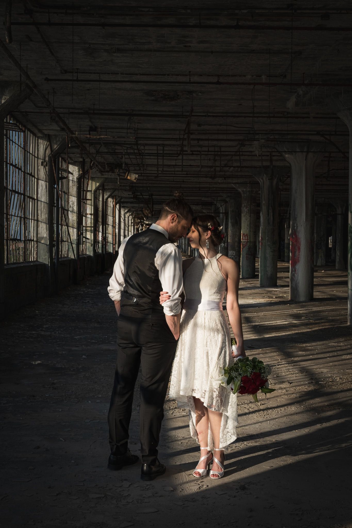 Bride and groom share a tender moment in a dramatic sunlit industrial hall with red roses.