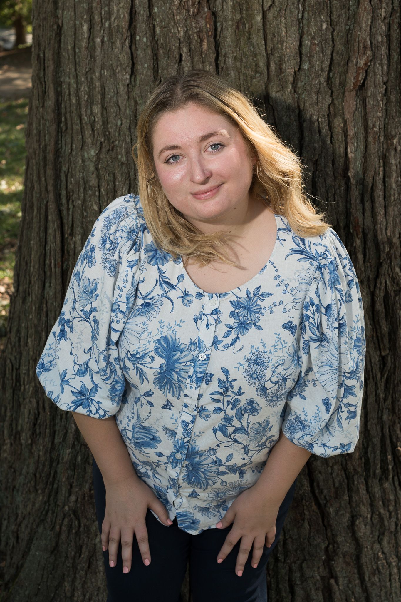 Young blonde woman in blue floral top leaning against large tree trunk outdoors.