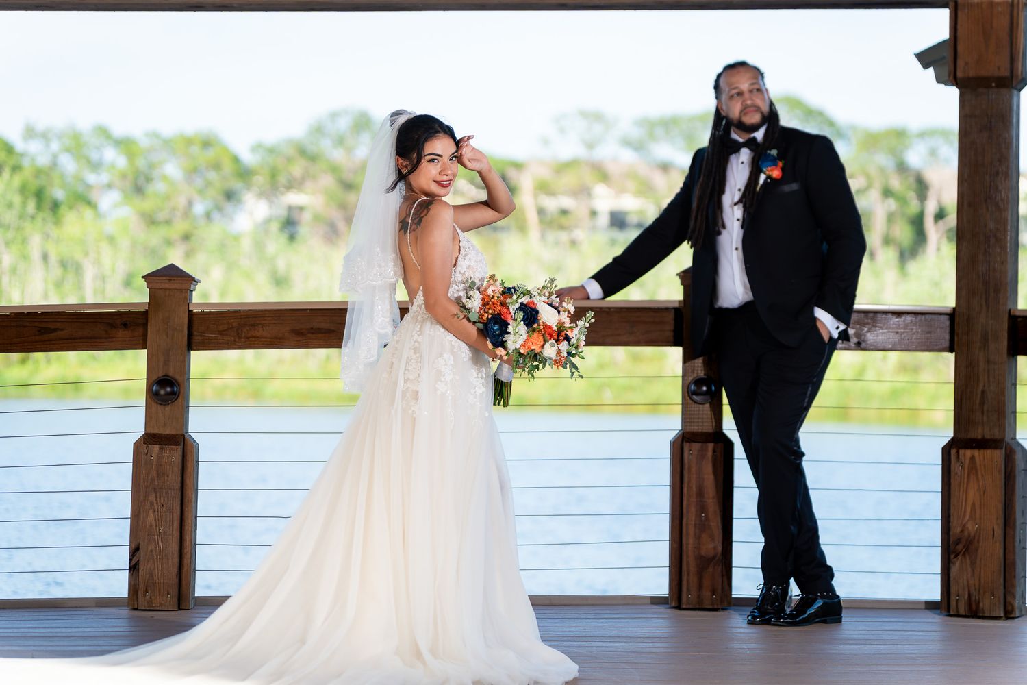 Bride in white gown holds bouquet while groom in black tuxedo stands on wooden dock by waterfront.