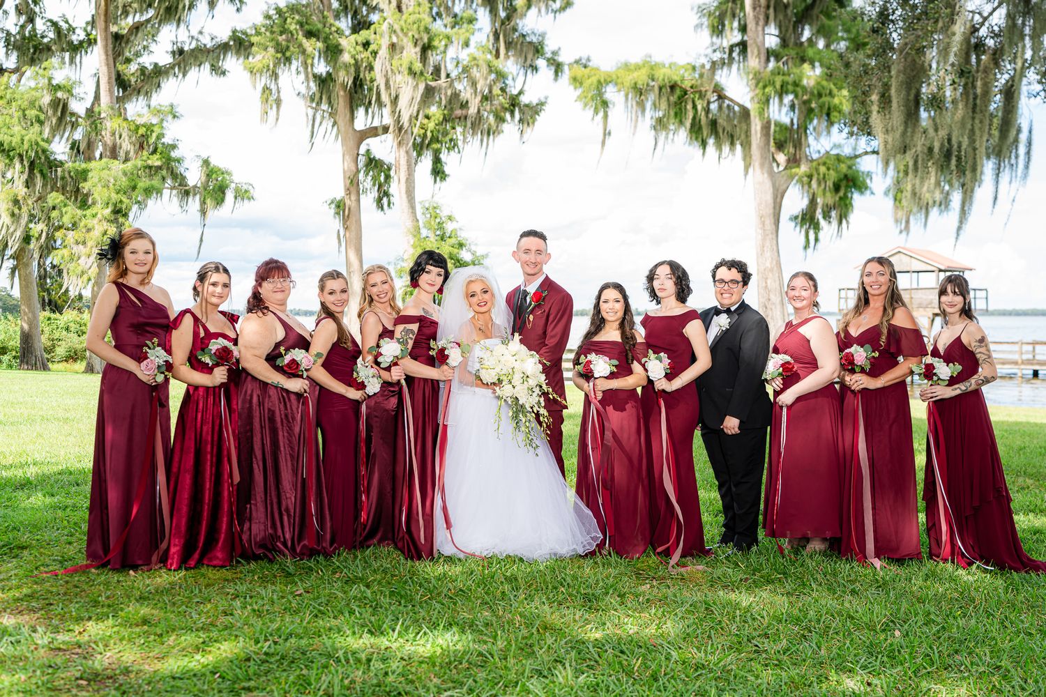 Wedding party poses with bride, groom, and bridesmaids in burgundy satin gowns under Spanish moss trees.