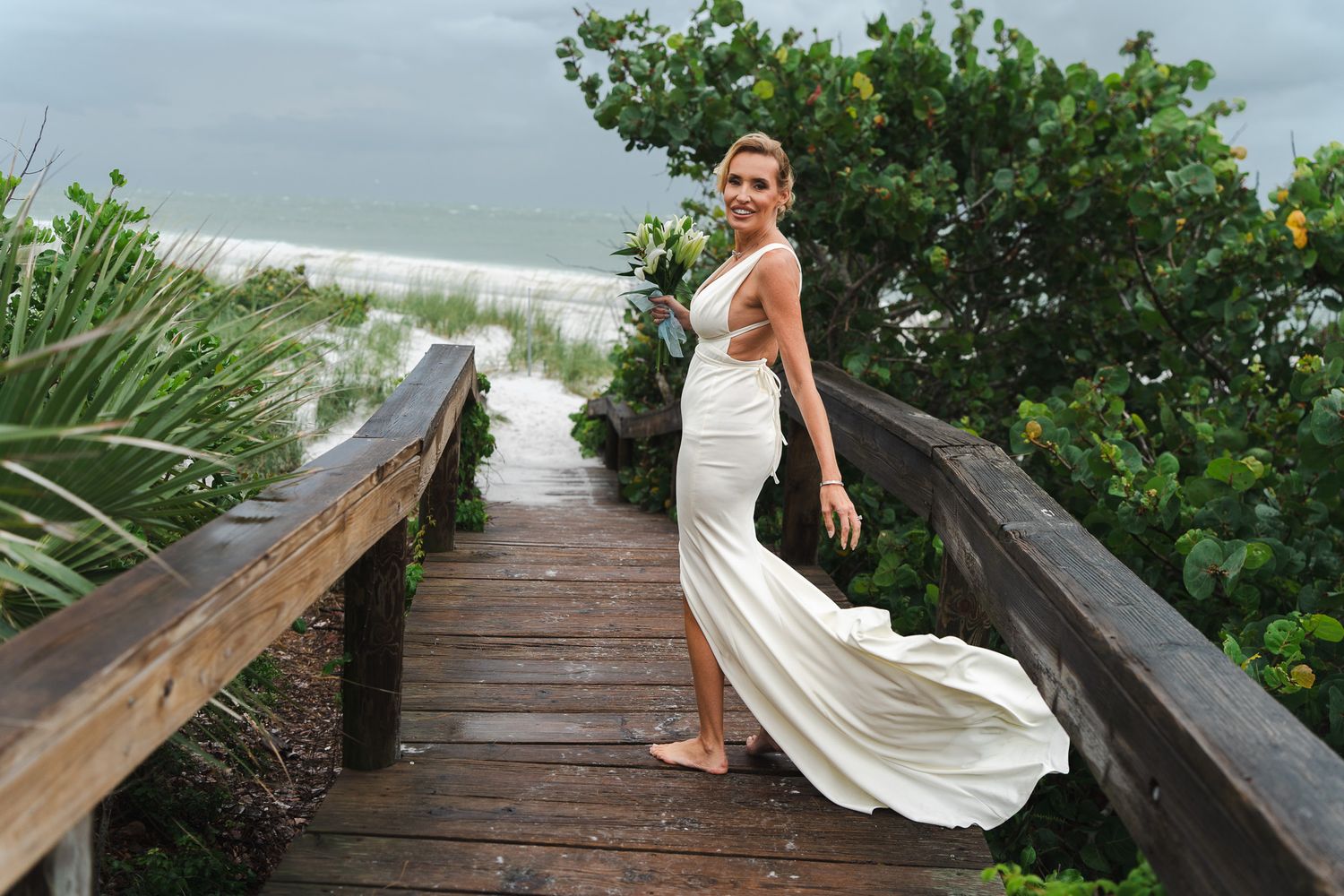 Bride in elegant white halter gown poses on wooden beach boardwalk surrounded by lush greenery.