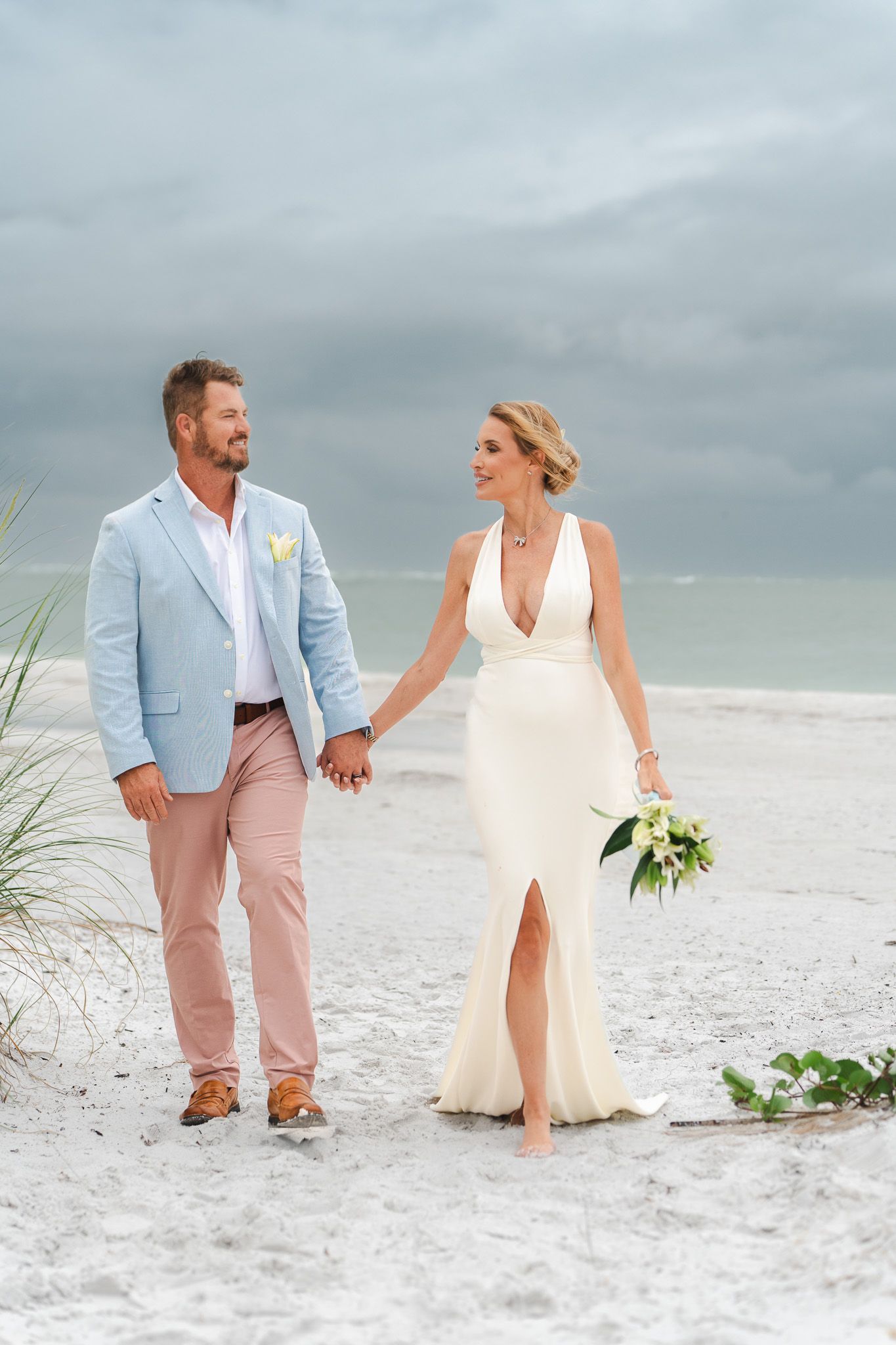 Smiling bride and groom holding hands walking on white sand beach with stormy sky in background.