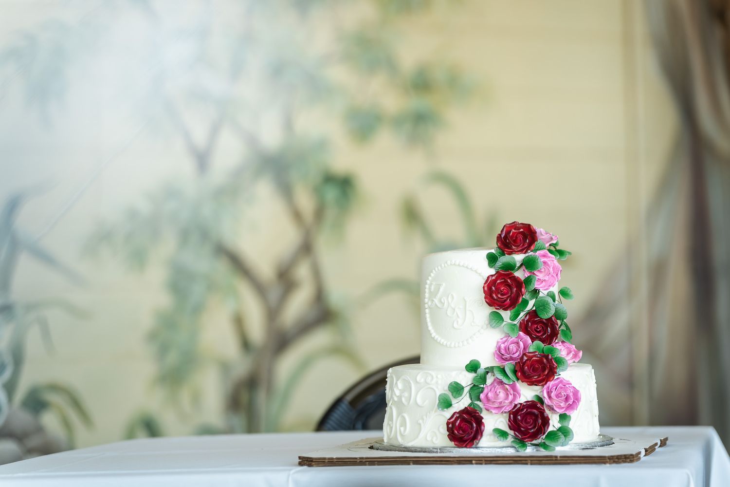 Two-tier white wedding cake adorned with red and pink sugar roses on decorative table.