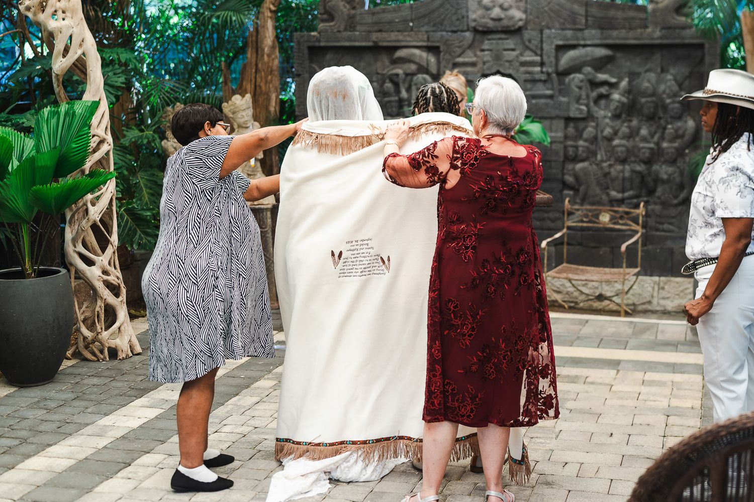 Guests help adjust bride's cape with embroidered text at outdoor wedding reception.
