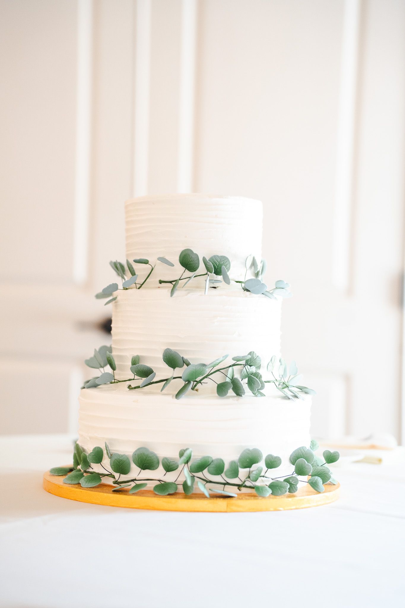 Three-tier white wedding cake decorated with fresh eucalyptus greenery on gold stand.