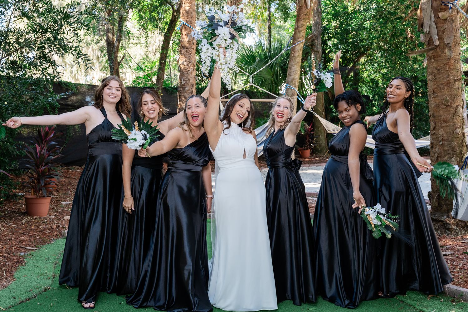 Bride in white dress poses with six bridesmaids in black satin gowns holding bouquets in lush garden.