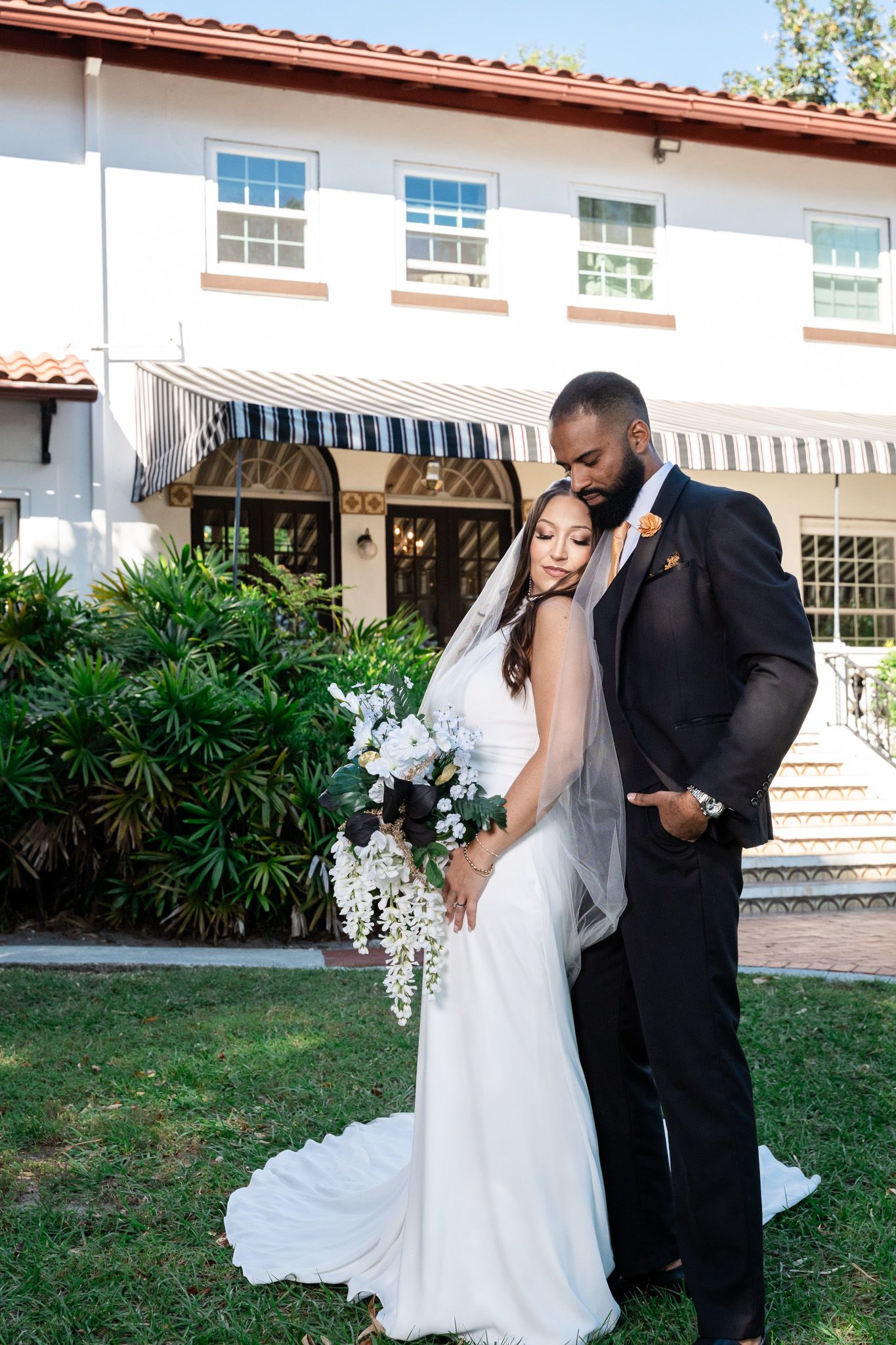 Bride with white bouquet and groom in black suit embrace tenderly in front of elegant venue.