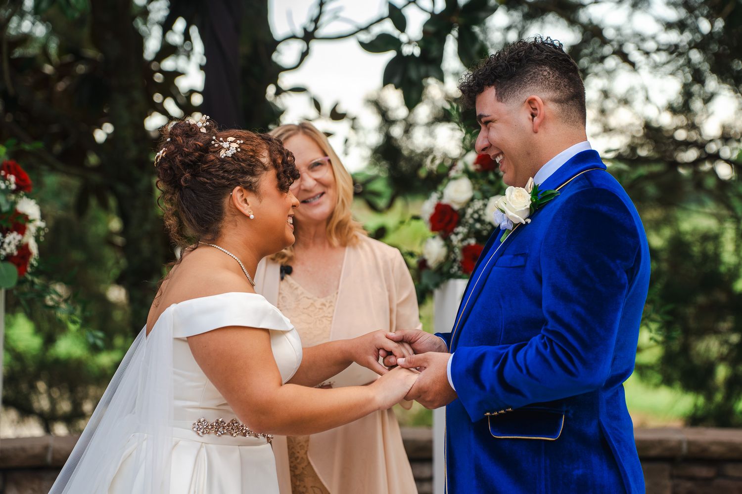 Bride places ring on grooms finger during outdoor wedding ceremony with officiant smiling.