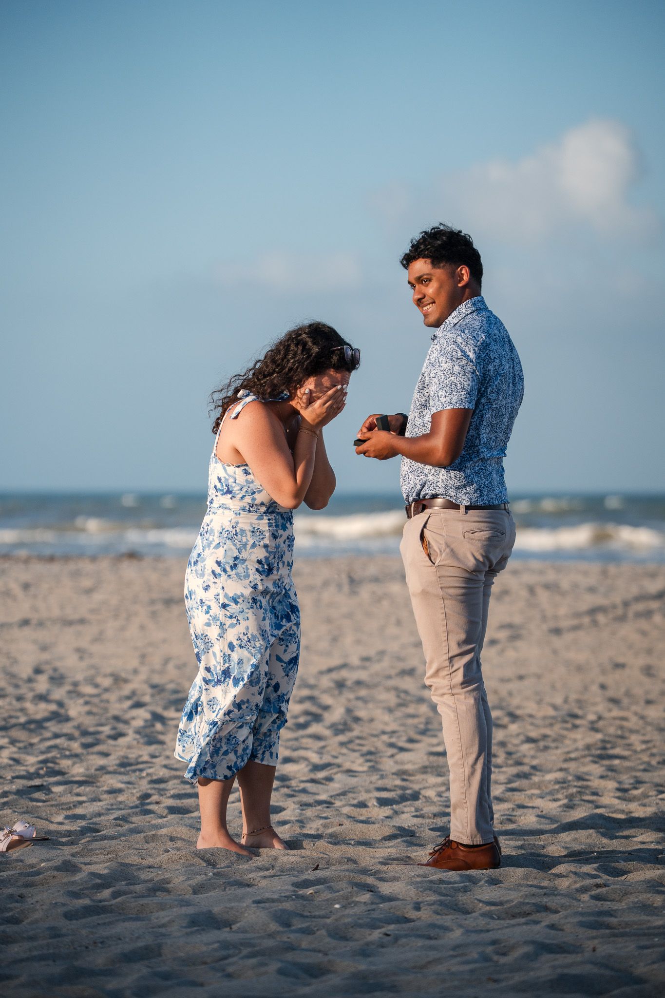 Man proposes to woman on sandy beach, she covers her face in surprise while he smiles holding a ring box.