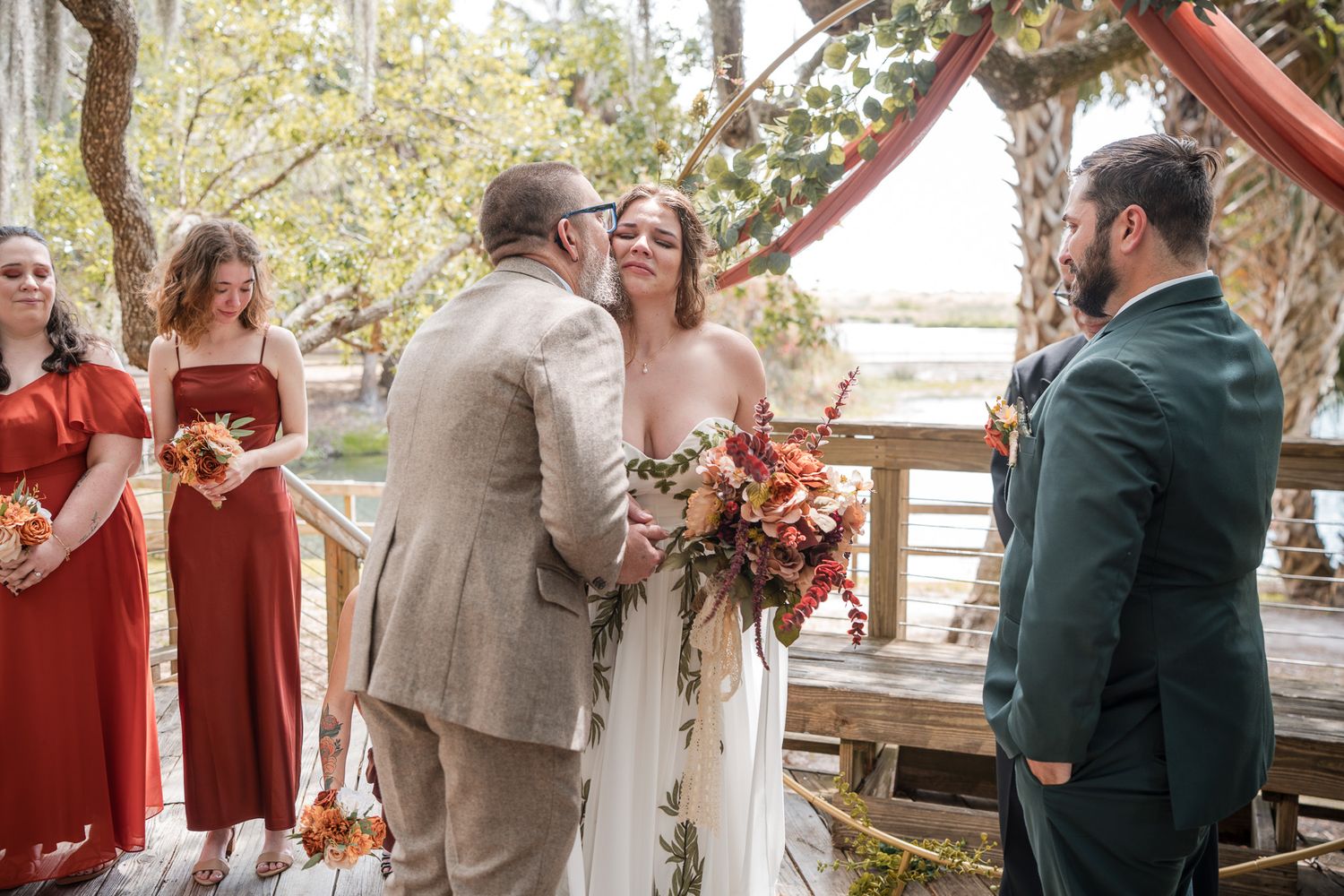 Bride and groom exchange vows on rustic wooden deck with red-dressed bridesmaids at outdoor wedding.