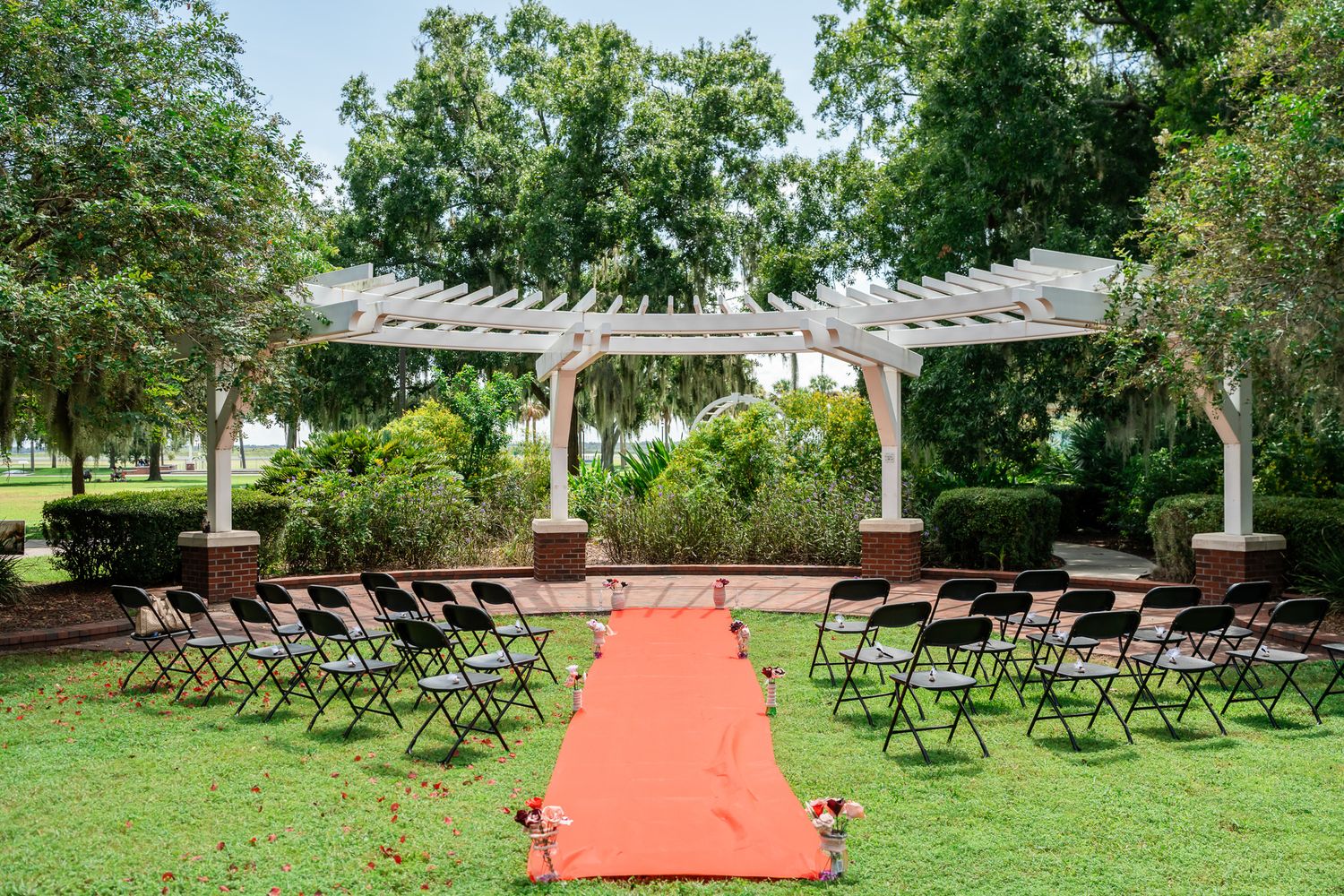 Outdoor wedding ceremony setup with red aisle runner, black chairs, and white pergola surrounded by lush green trees.