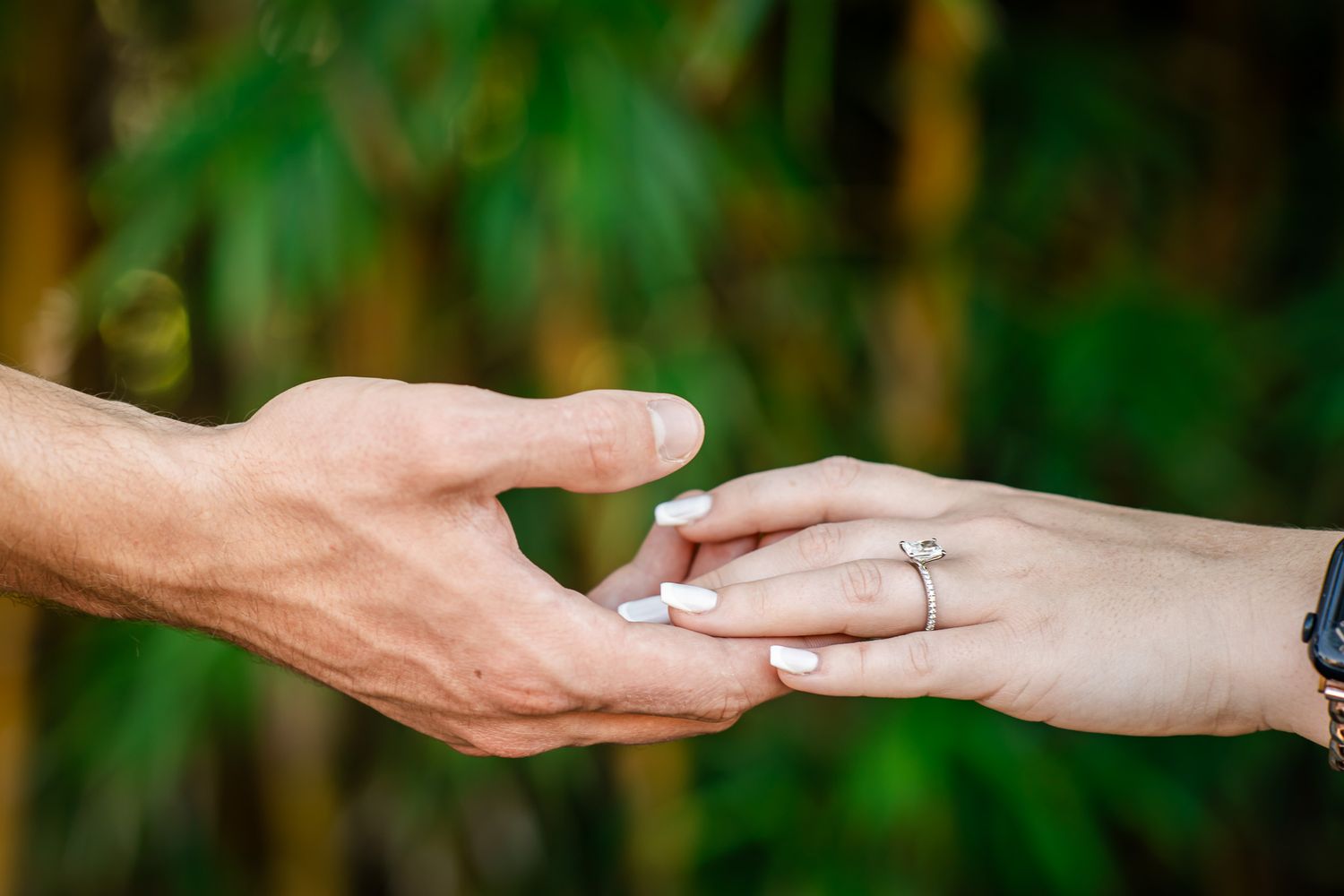 Close-up of a couple gently touching hands outdoors, woman wearing an engagement ring with bamboo background.