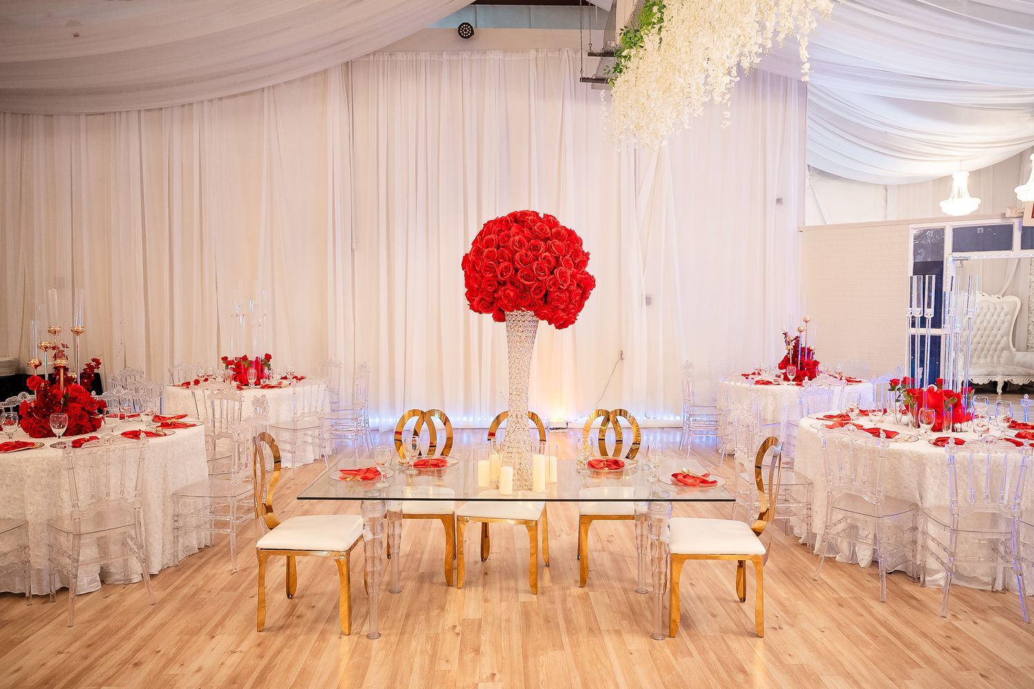 Elegant wedding reception venue with red rose centerpieces, gold chairs, glass tables, and white draping.