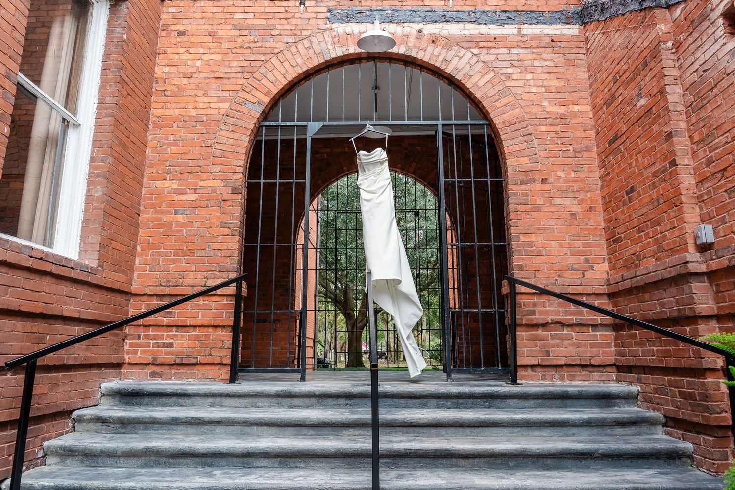 White wedding dress hanging in arched iron gate doorway of a red brick wedding venue.