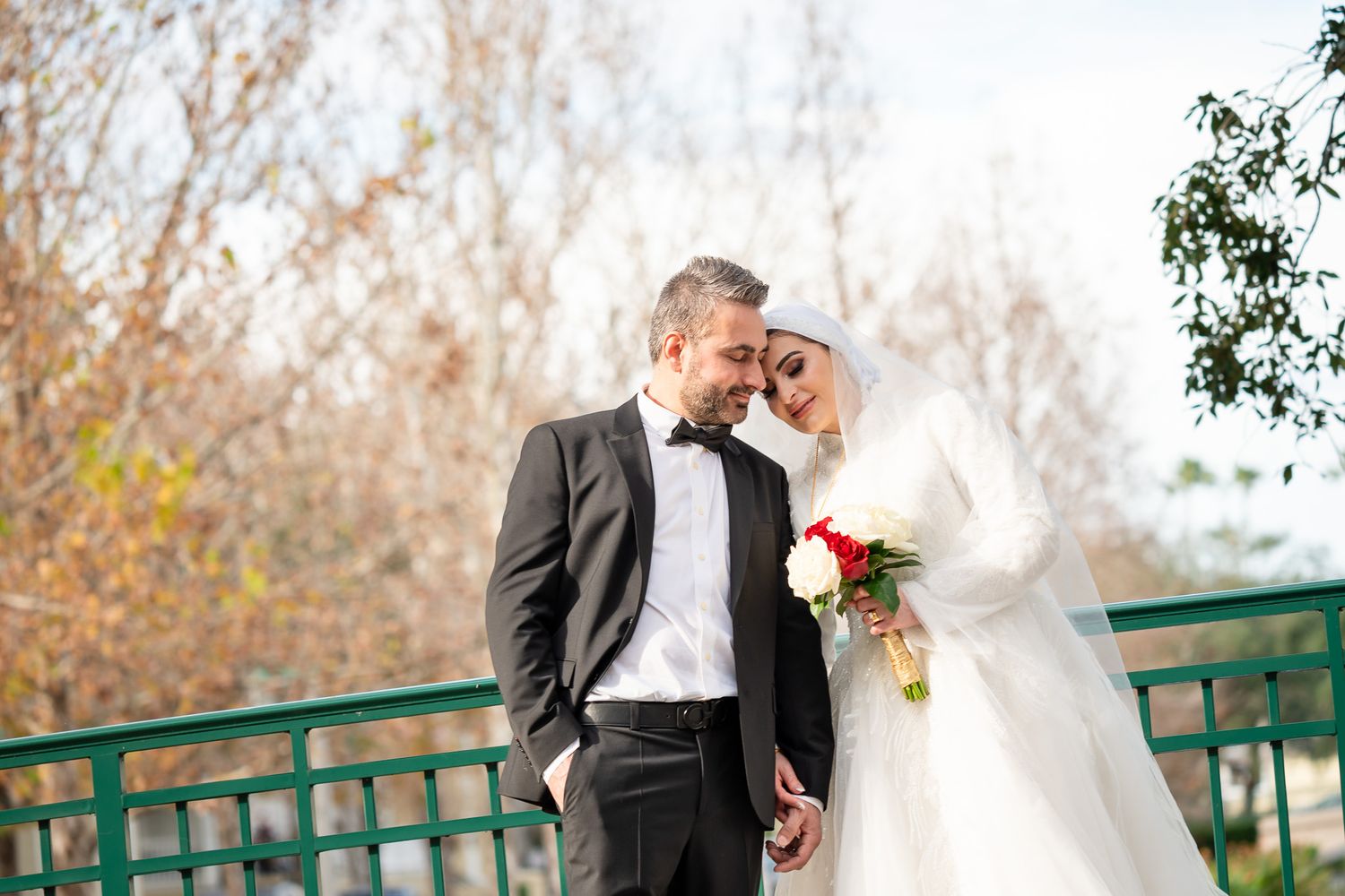 Bride and groom share a tender moment on a bridge surrounded by autumn trees in soft natural light.