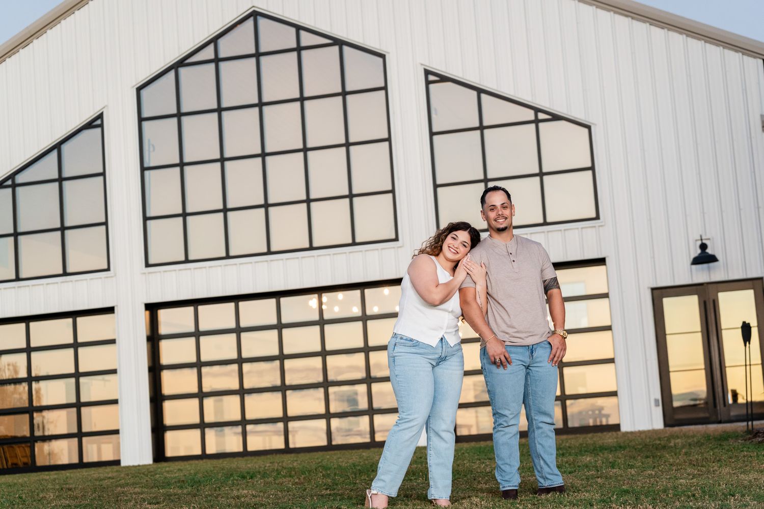 Smiling engaged couple poses in front of large white barn at sunset.