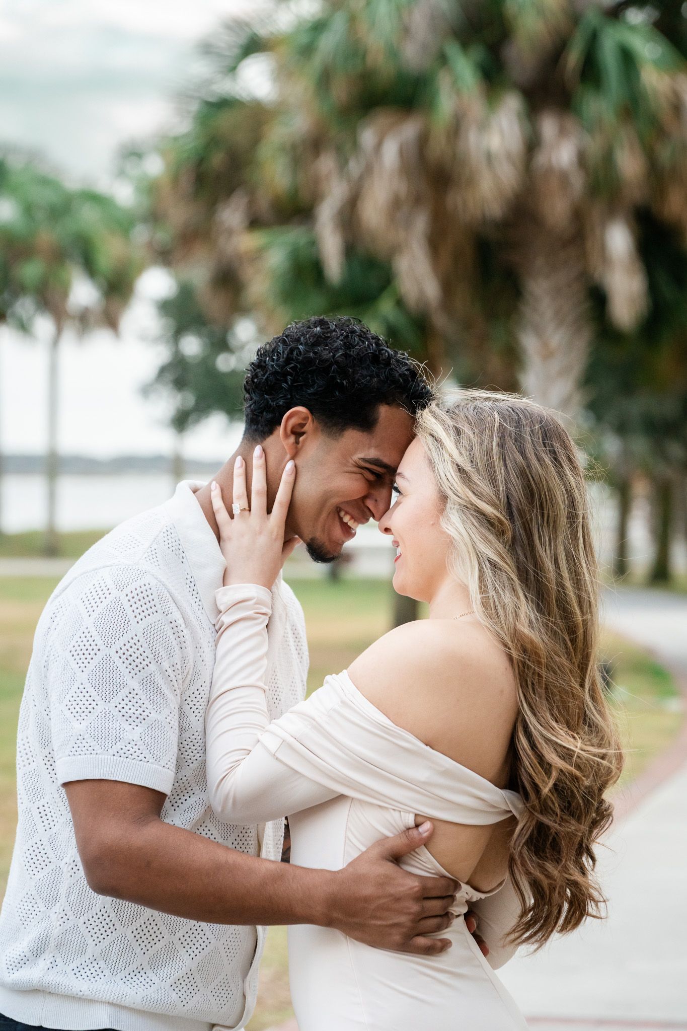Smiling interracial couple shares an intimate moment touching foreheads near tropical palm trees.