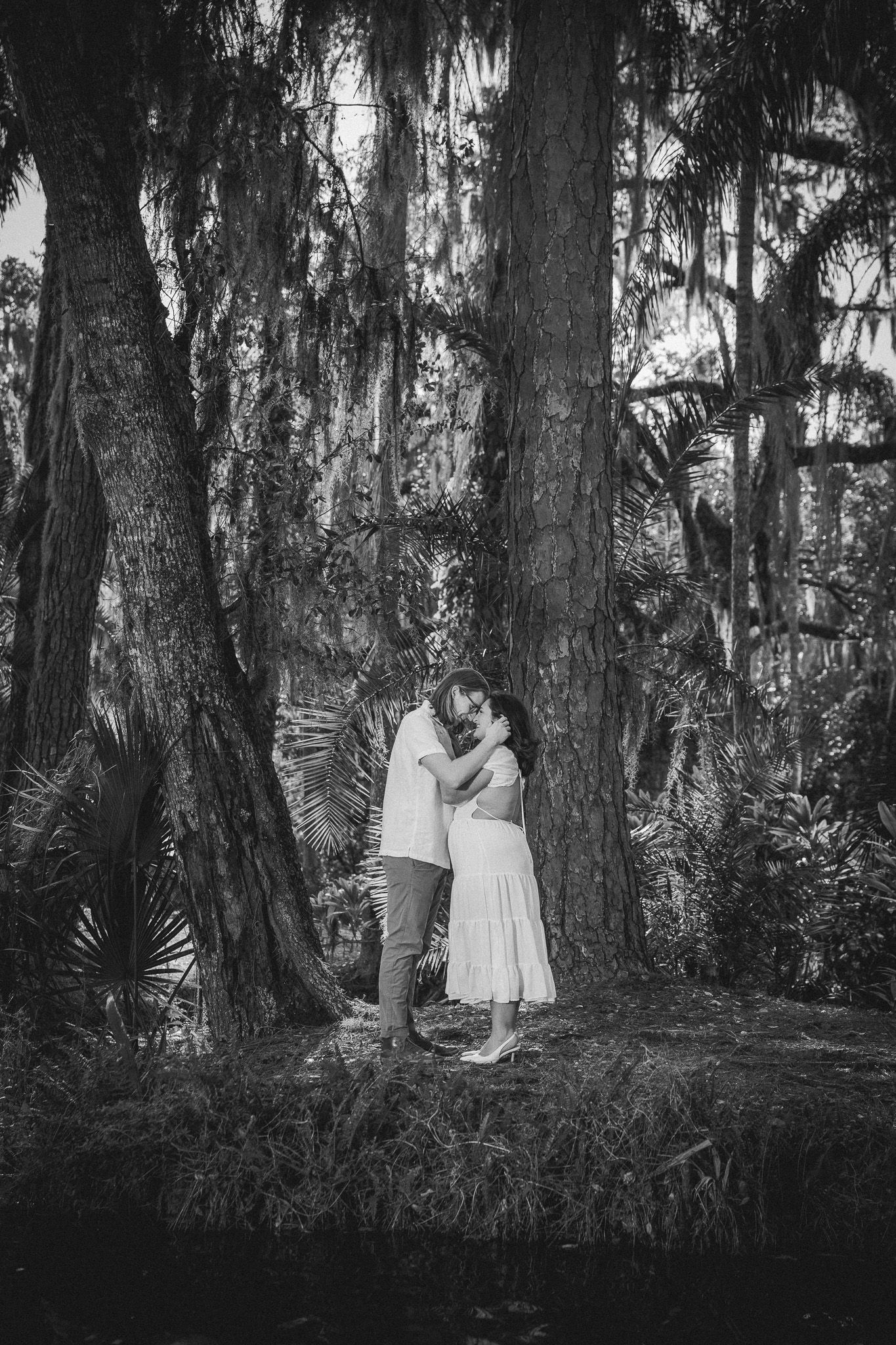 Black and white photo of a couple kissing under large moss-draped trees in a lush forest setting.