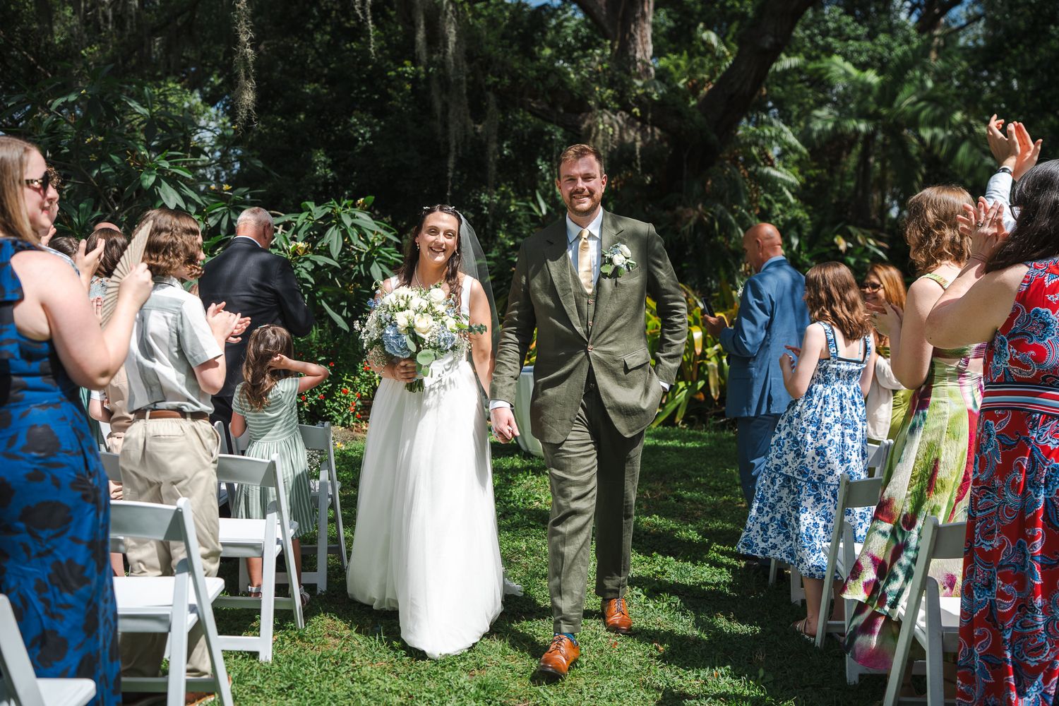 Joyful bride and groom walk back down the aisle after their outdoor garden wedding ceremony as guests celebrate.