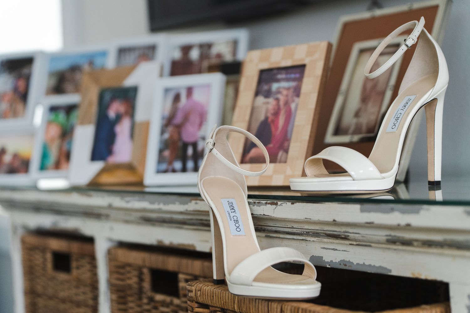 White Jimmy Choo strappy heeled sandals displayed on a rustic shelf surrounded by framed family photographs.