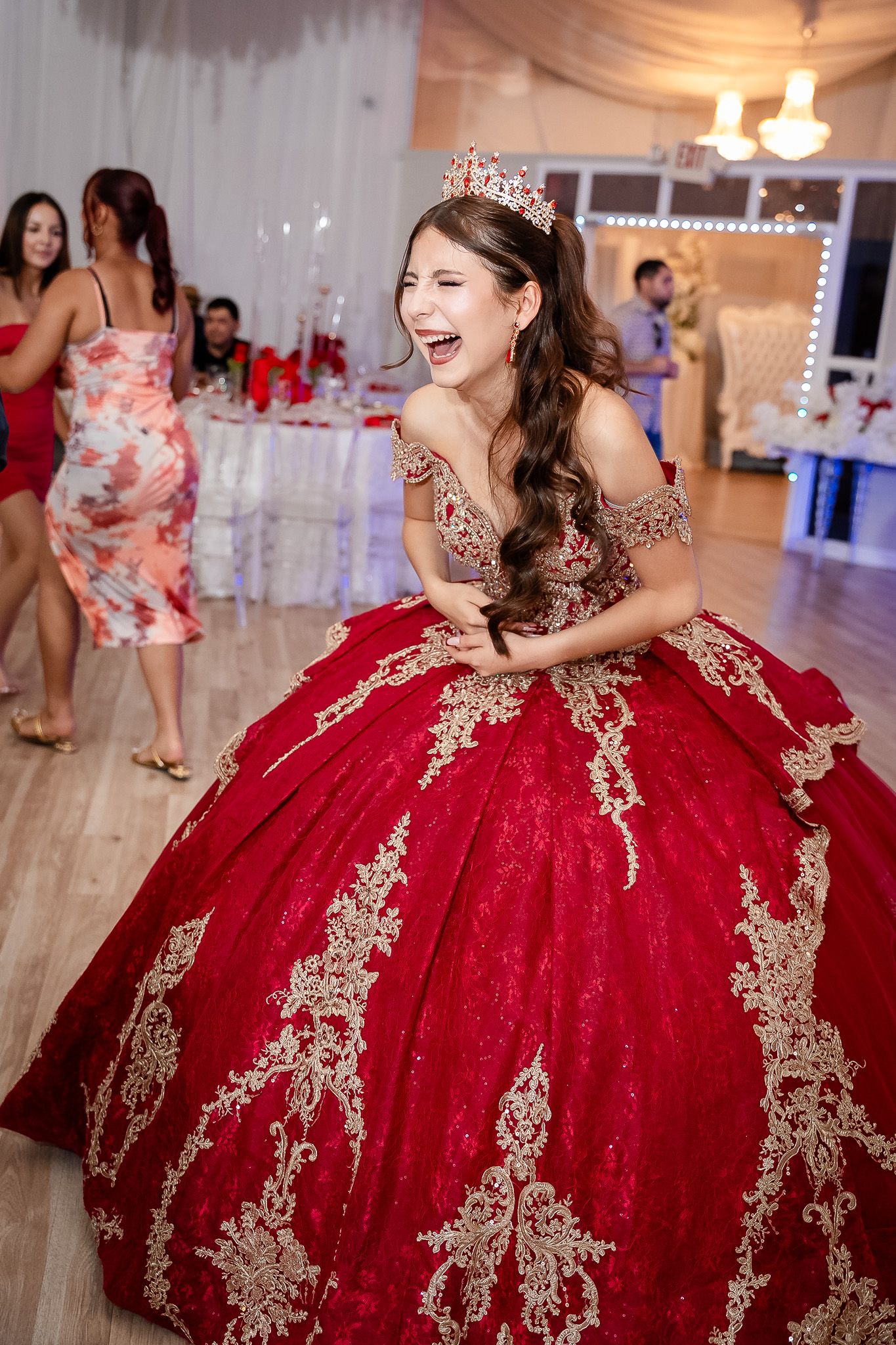 Joyful quinceañera girl laughing in a stunning red and gold ballgown with tiara at her party.