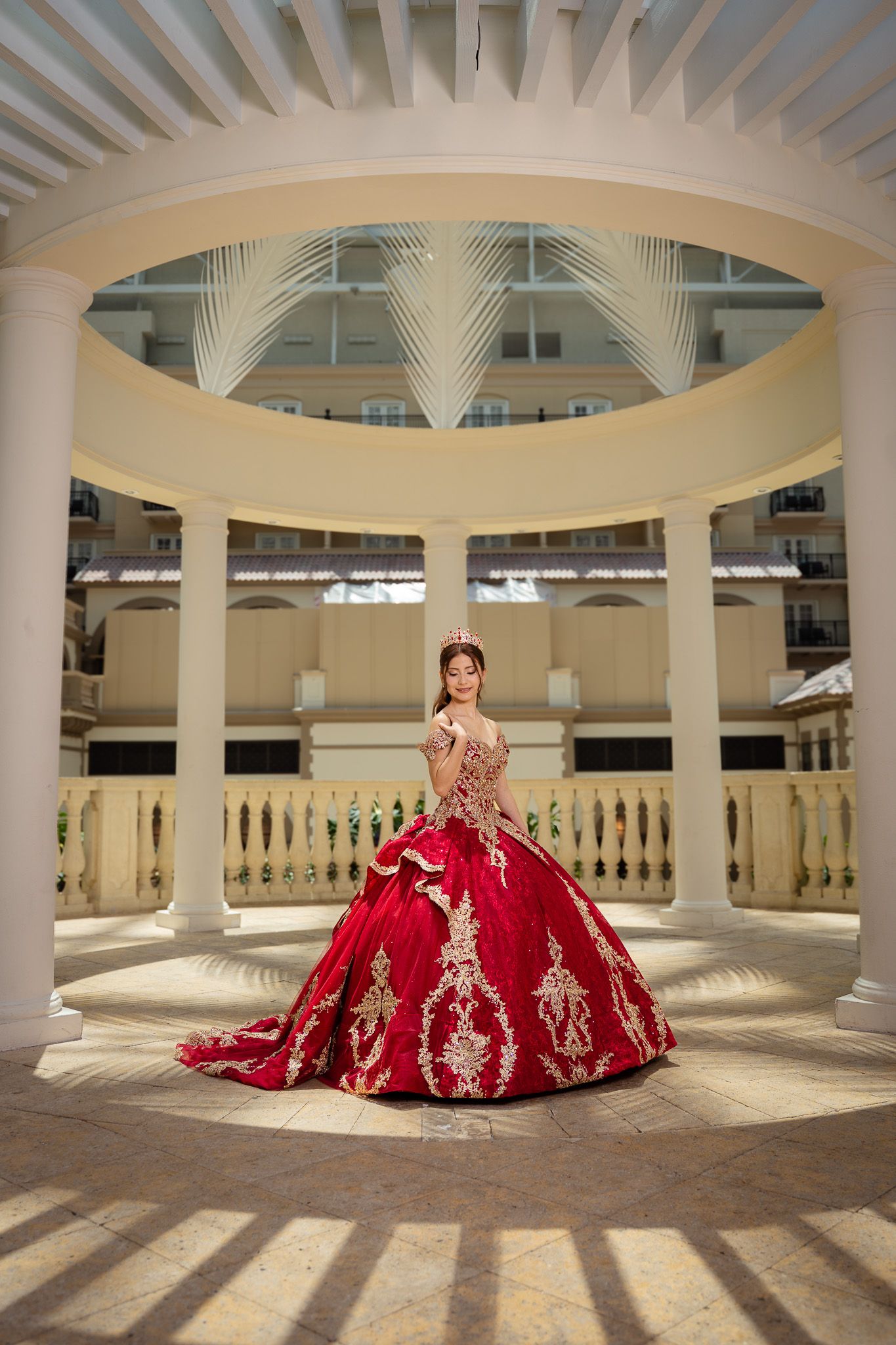 Young woman in a stunning red and gold ball gown quinceañera dress poses in an elegant white columned rotunda.