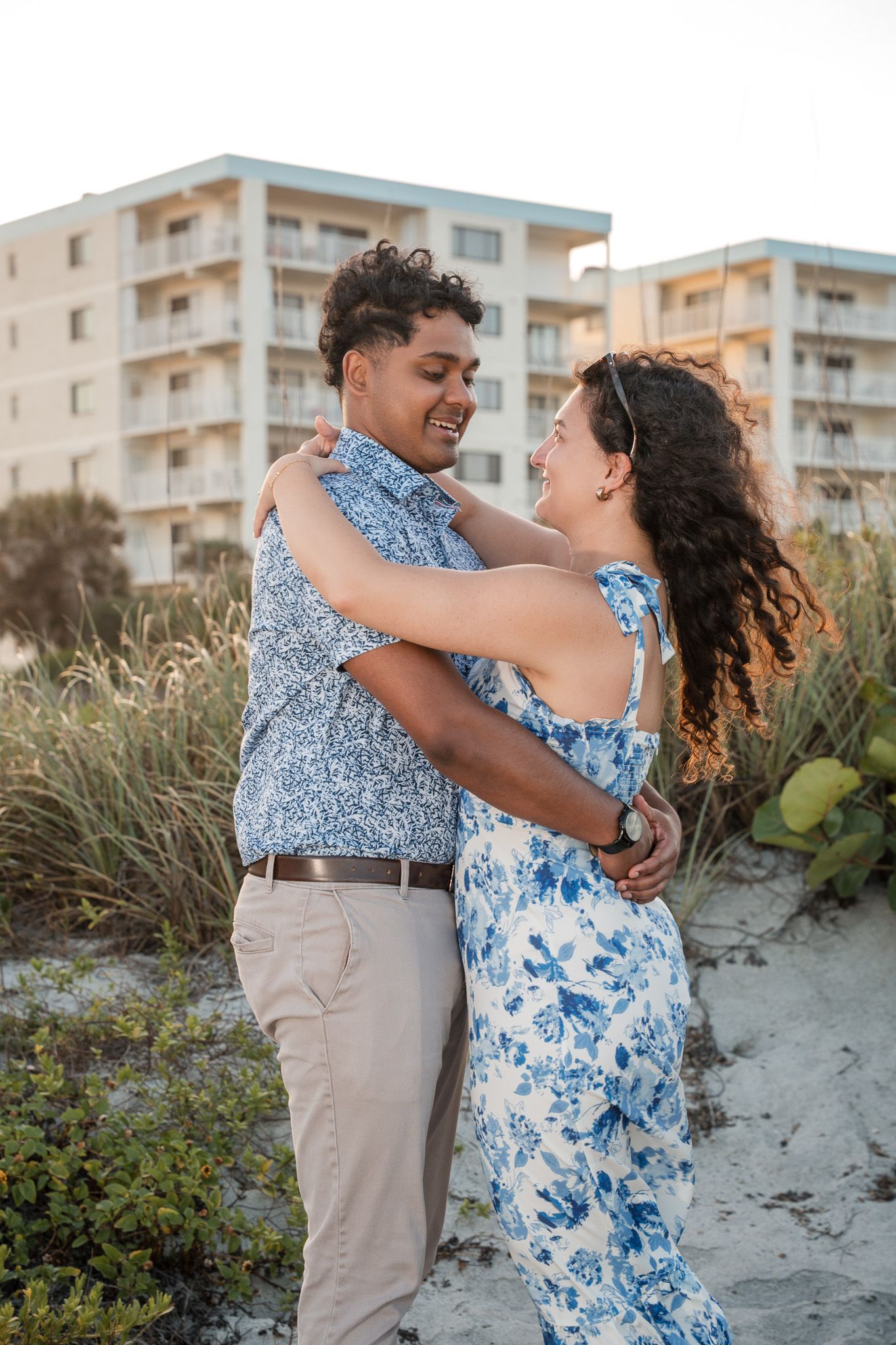 Happy interracial couple embracing and smiling at each other on sandy beach with resort building behind.