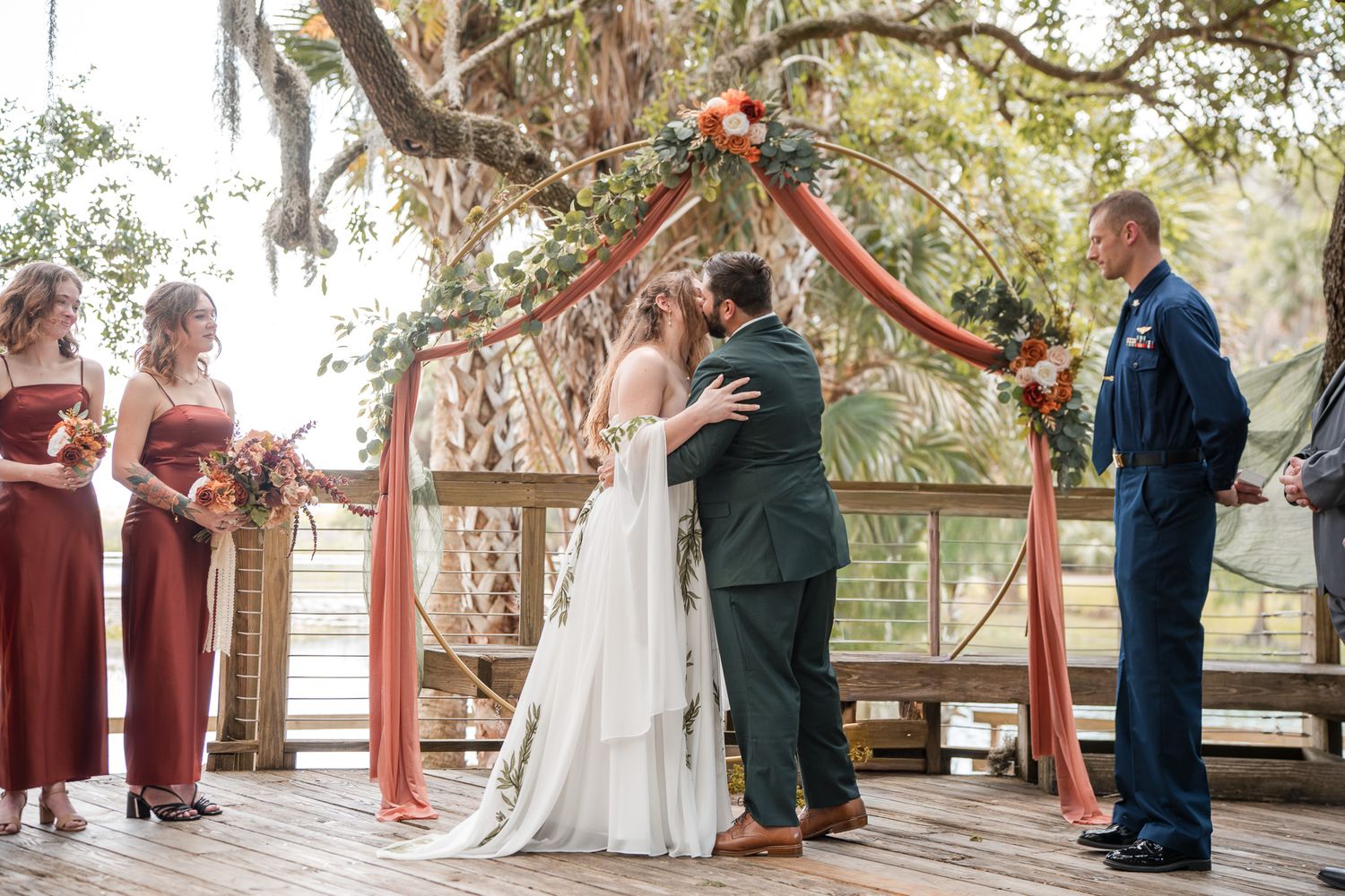 Bride and groom share first kiss at outdoor wedding ceremony on wooden deck with floral arch and bridesmaids in rust dresses.