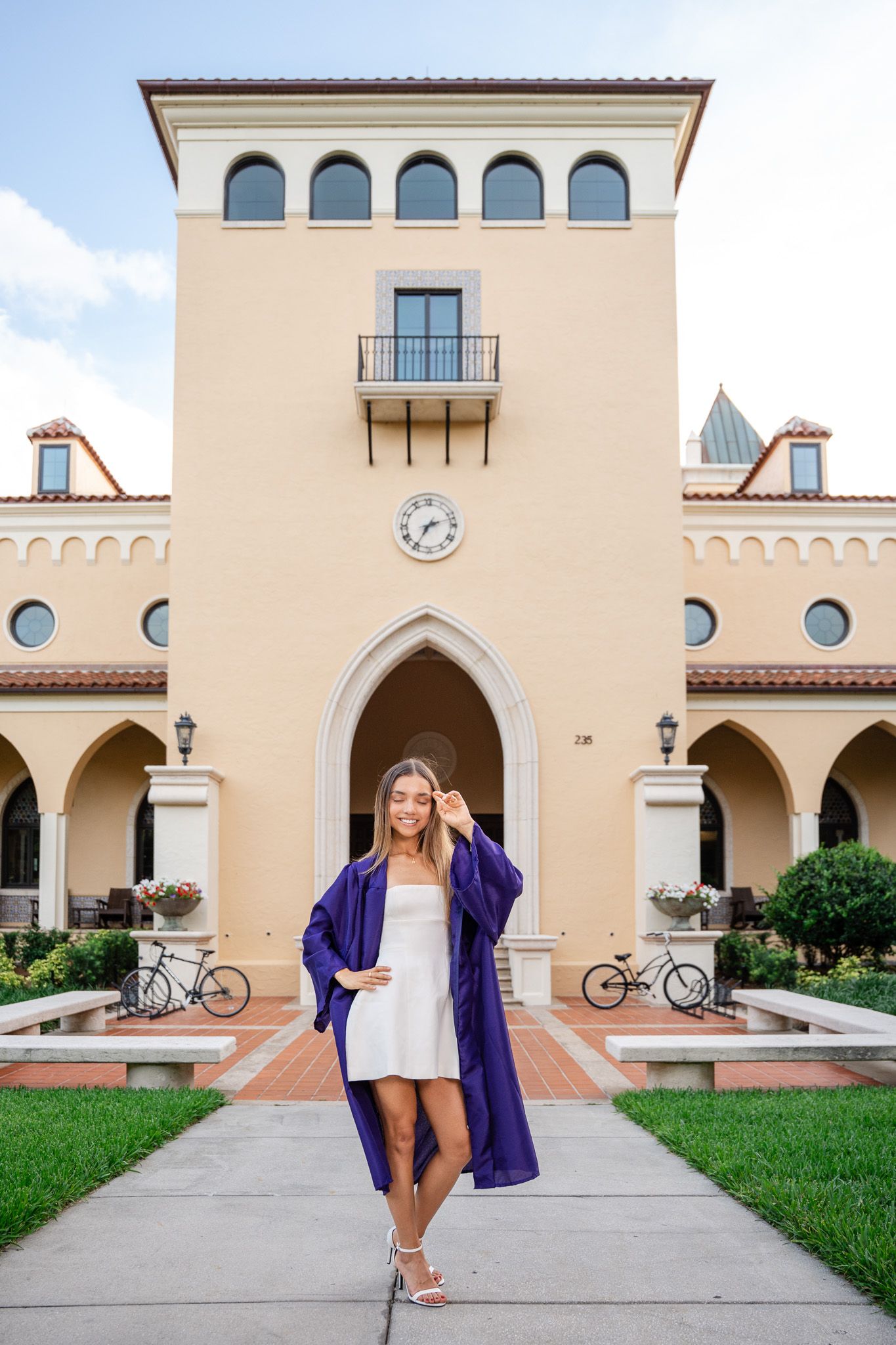 Graduate in purple gown adjusting cap while walking in front of elegant university building.