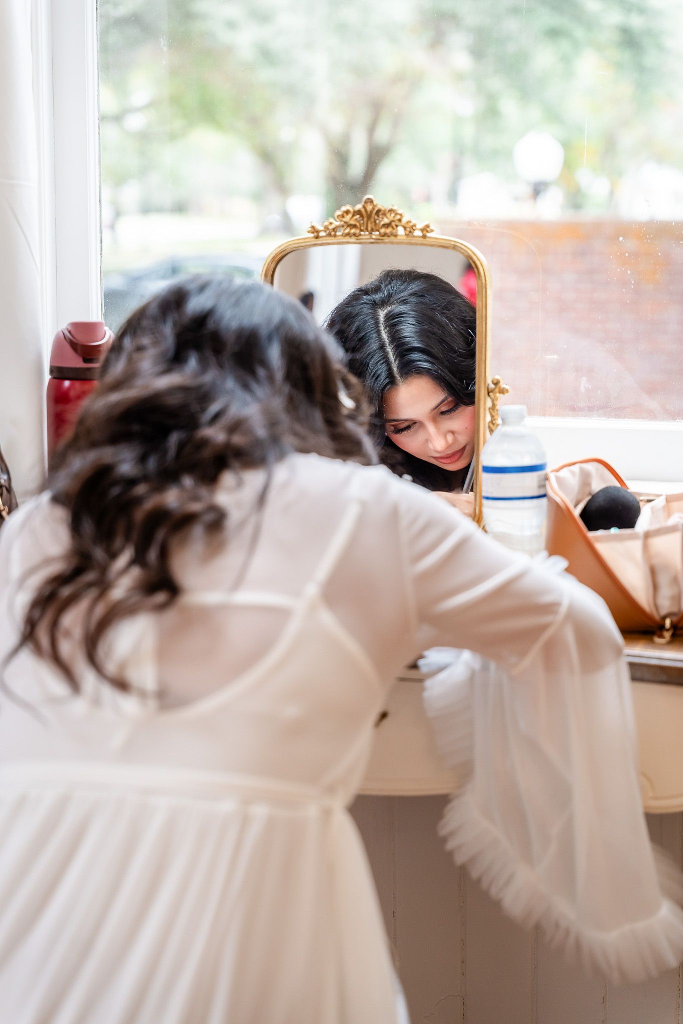 Bride in white robe leans toward gold-framed mirror while getting ready for her wedding.