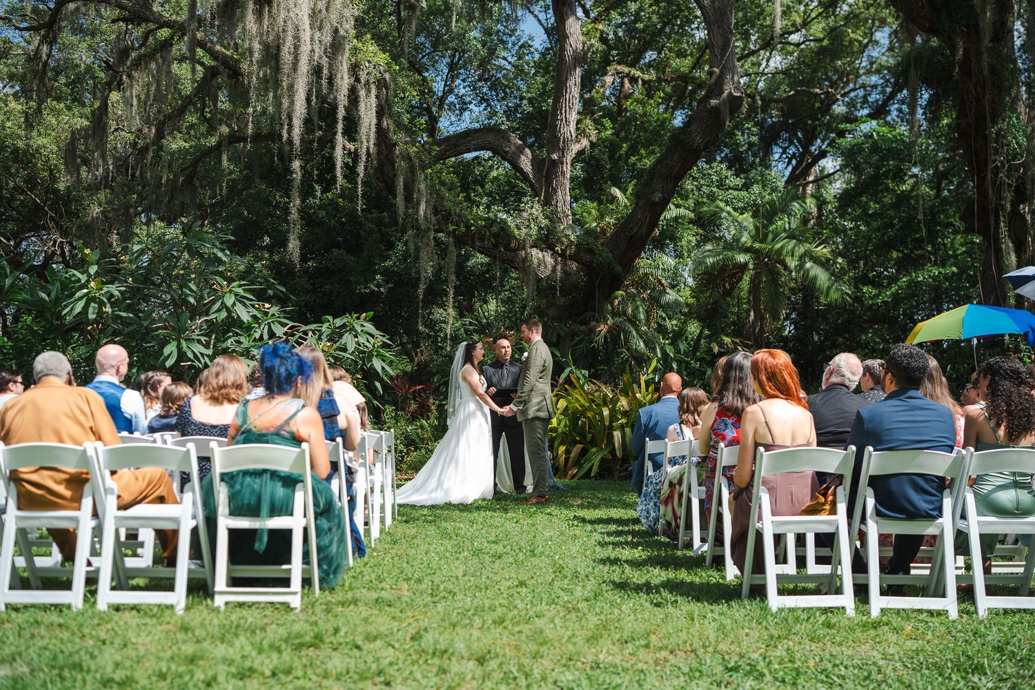 Outdoor garden wedding ceremony with guests seated in white chairs beneath moss-draped oak trees.