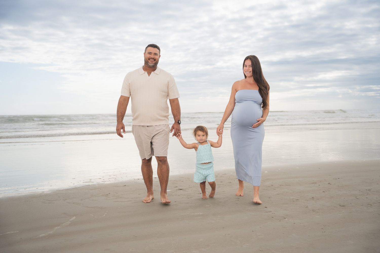 Pregnant woman in blue dress and man walk hand-in-hand with toddler on sandy beach.