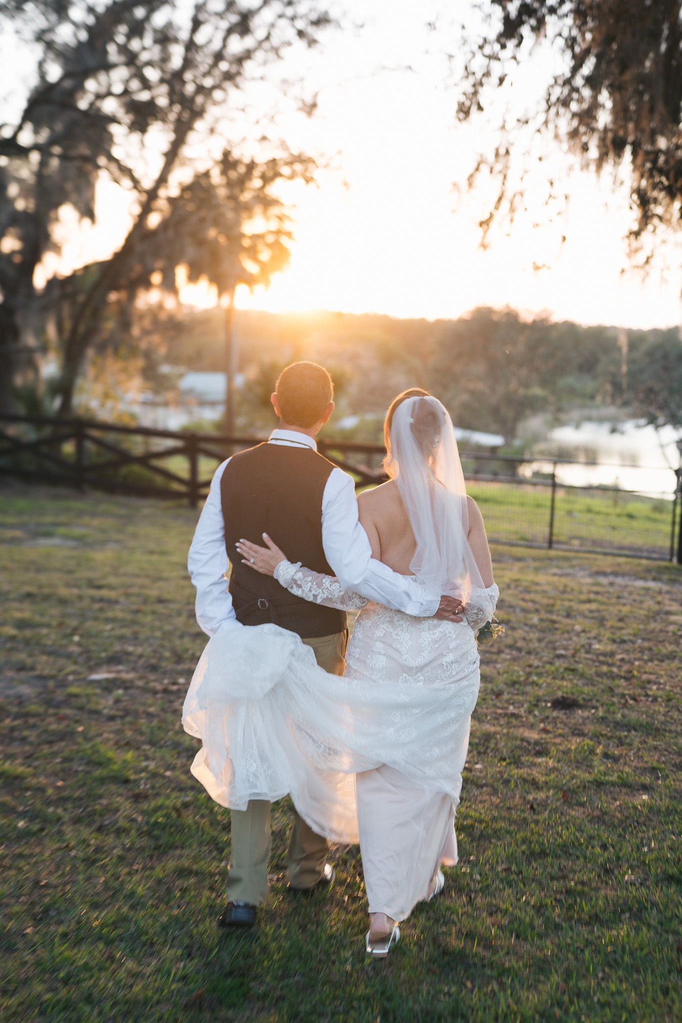 Bride and groom walk hand in hand toward stunning golden sunset at waterfront wedding.