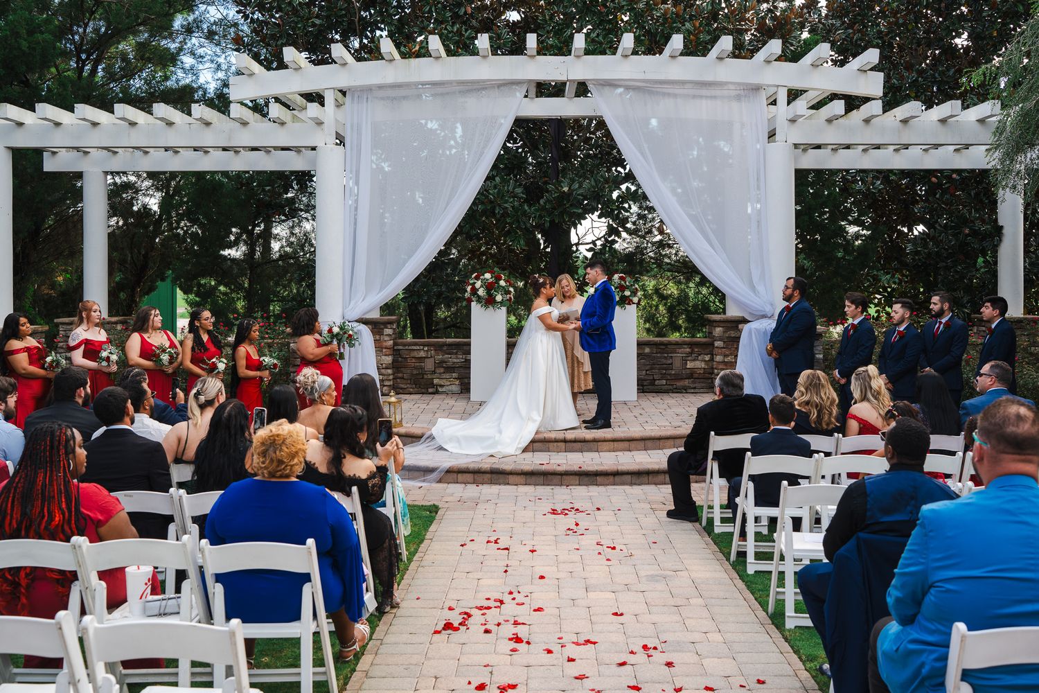 Wide shot of outdoor wedding ceremony under white pergola with red and white floral decor.