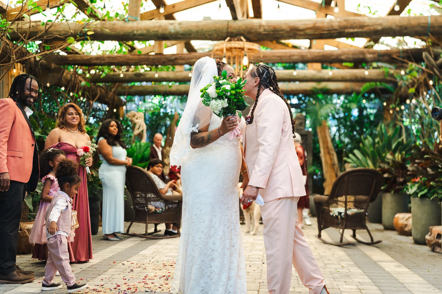 Brides share intimate moment at lush garden wedding ceremony under wooden pergola with guests watching.