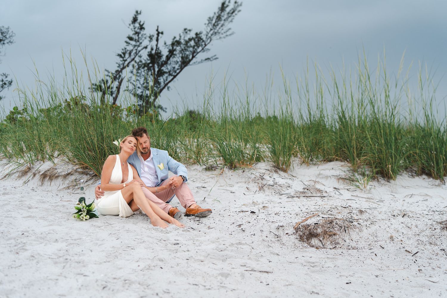 Newlywed couple sitting together on white sand dunes surrounded by sea grass on a cloudy beach day.