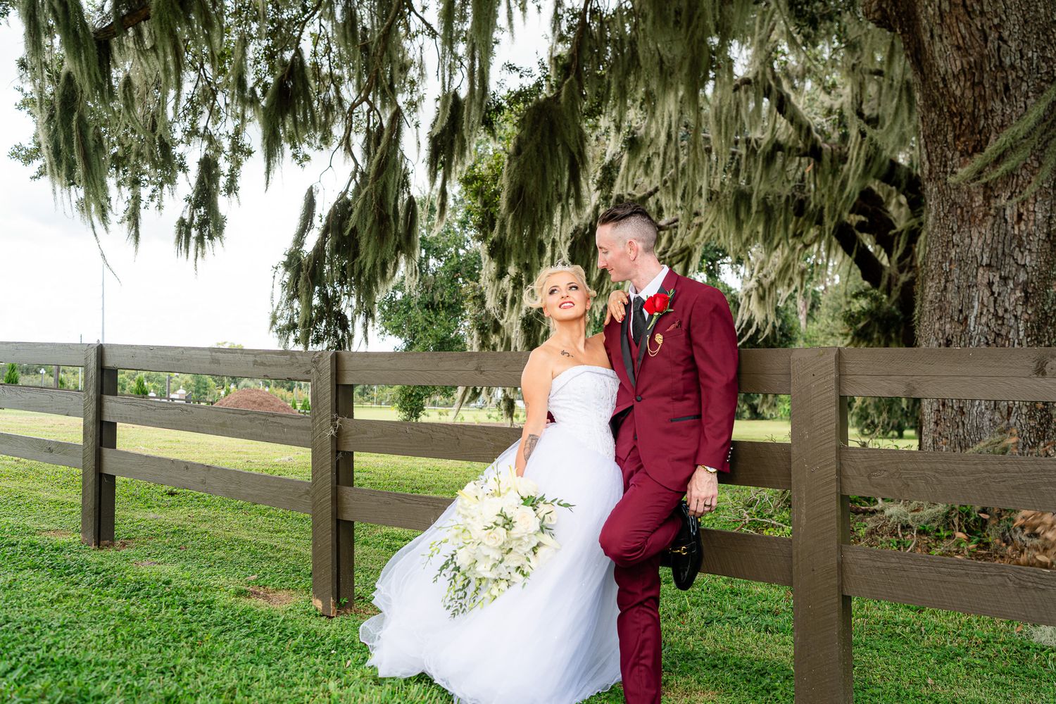 Bride and groom share a romantic moment leaning against wooden fence under Spanish moss-draped oak tree.