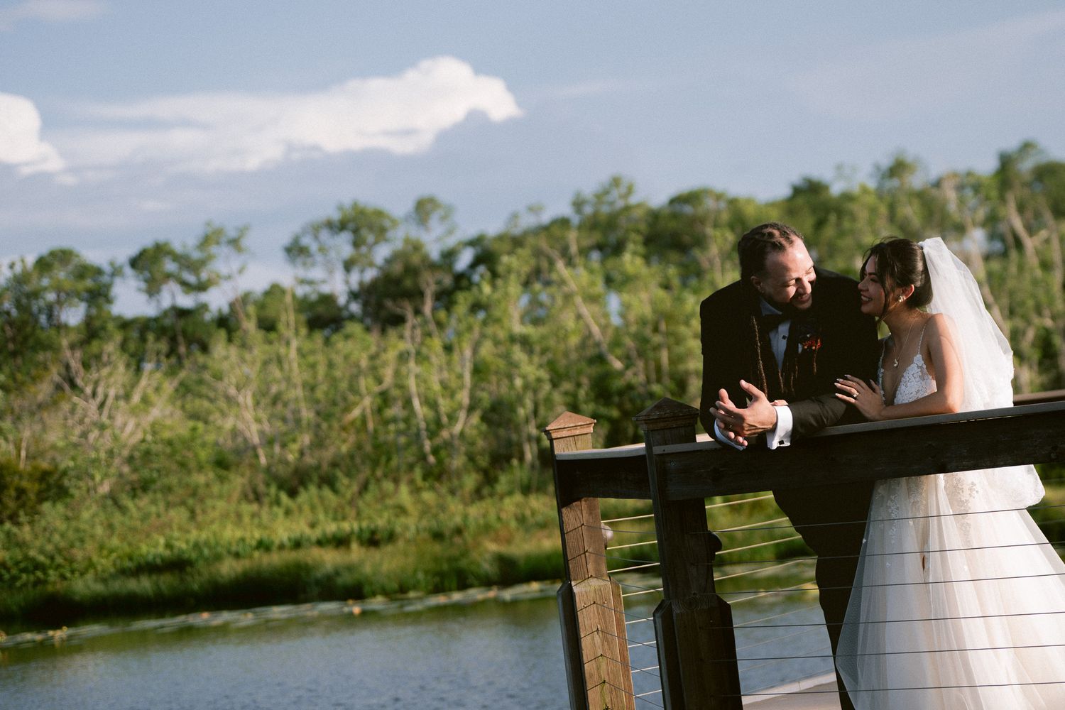 Bride and groom lean on wooden railing of waterfront dock sharing a romantic moment.