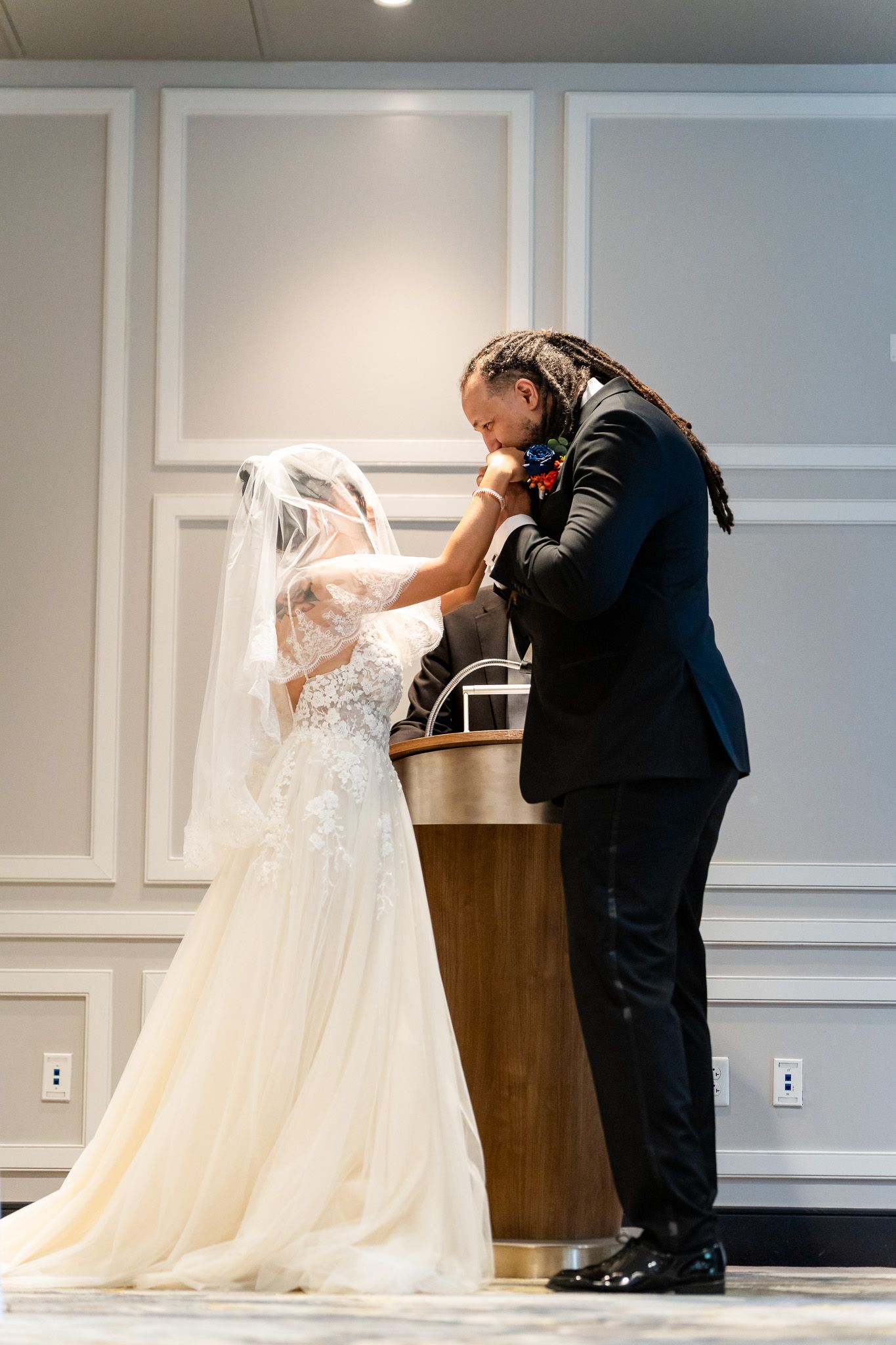 Groom lifting bride's veil at wedding altar in elegant white paneled room.