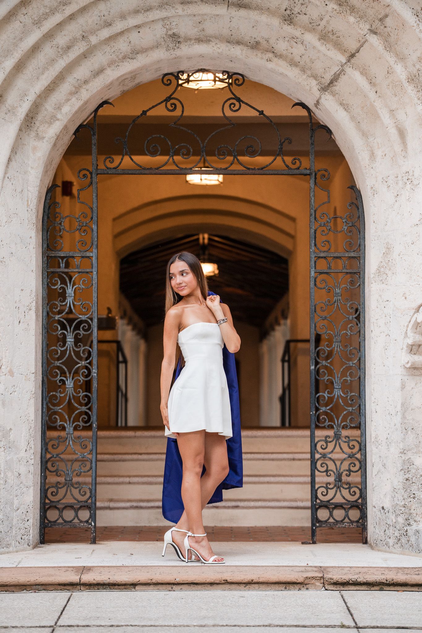 Young woman in white strapless dress and heels poses under ornate iron arch gateway of elegant stone building.