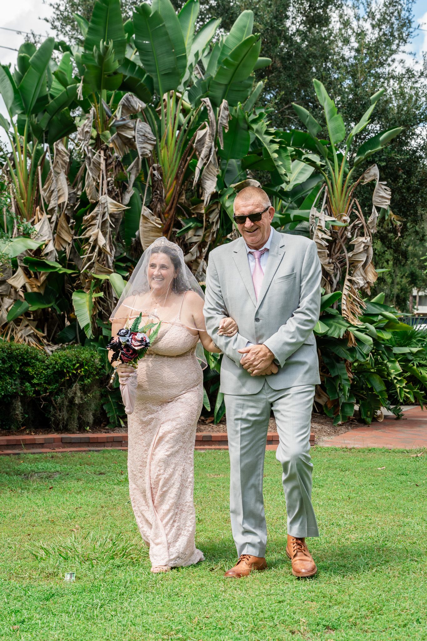 Bride in white gown and veil walks with groom in gray suit outdoors, tropical greenery background.