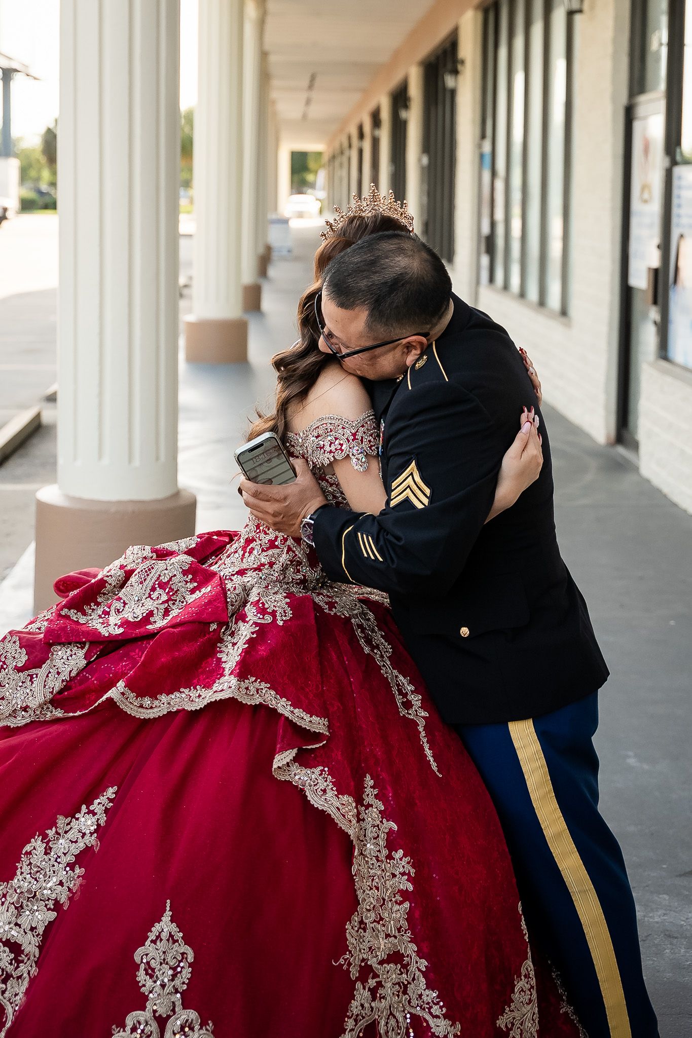 Soldier in dress uniform embraces young woman in red gold quinceañera ball gown outdoors.