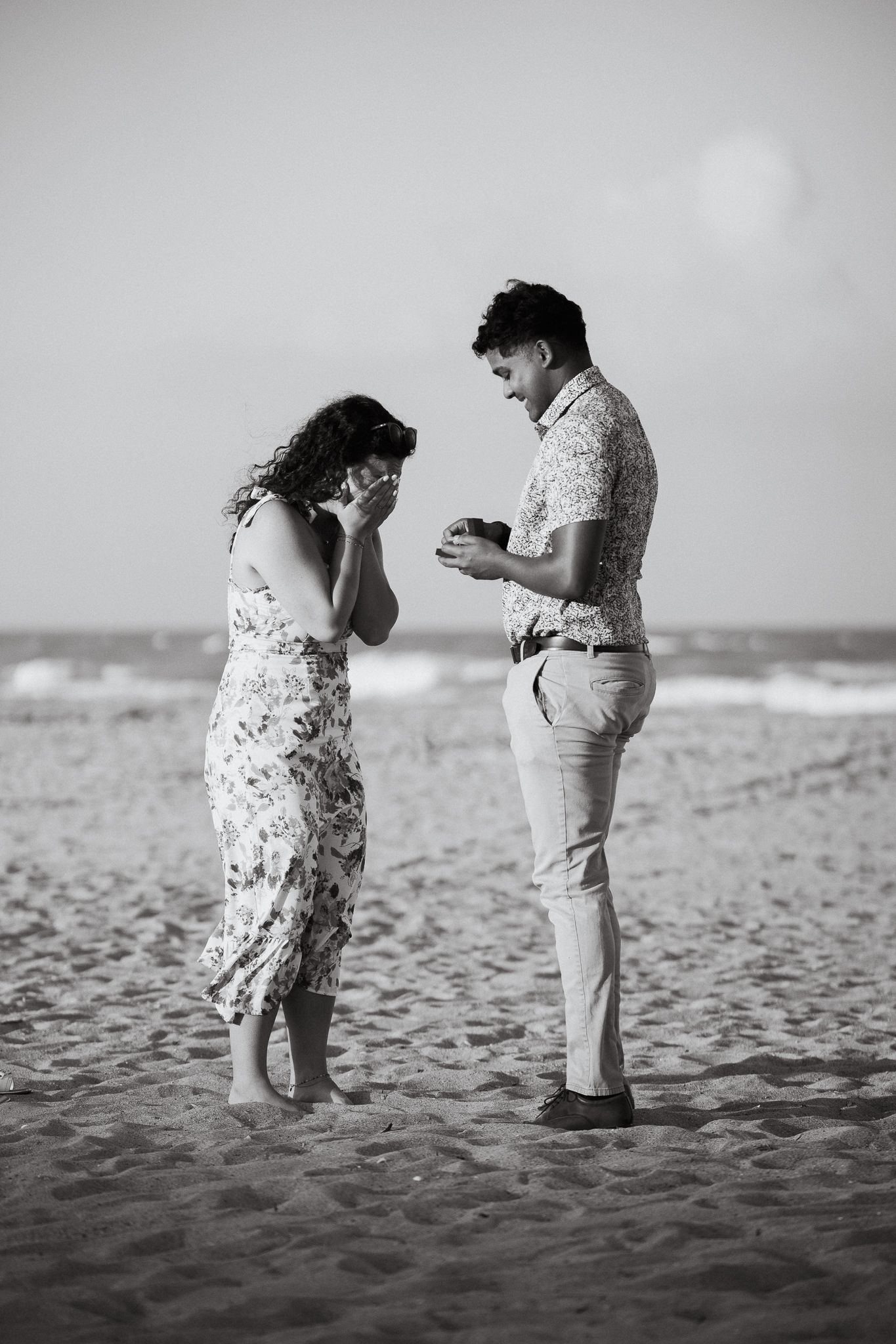 Black and white photo of a man proposing to a woman on a sandy beach with ocean waves in the background.