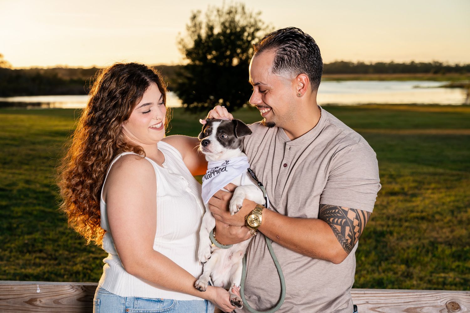 Smiling couple holding their small dog together near a wooden bench at a sunny outdoor location.