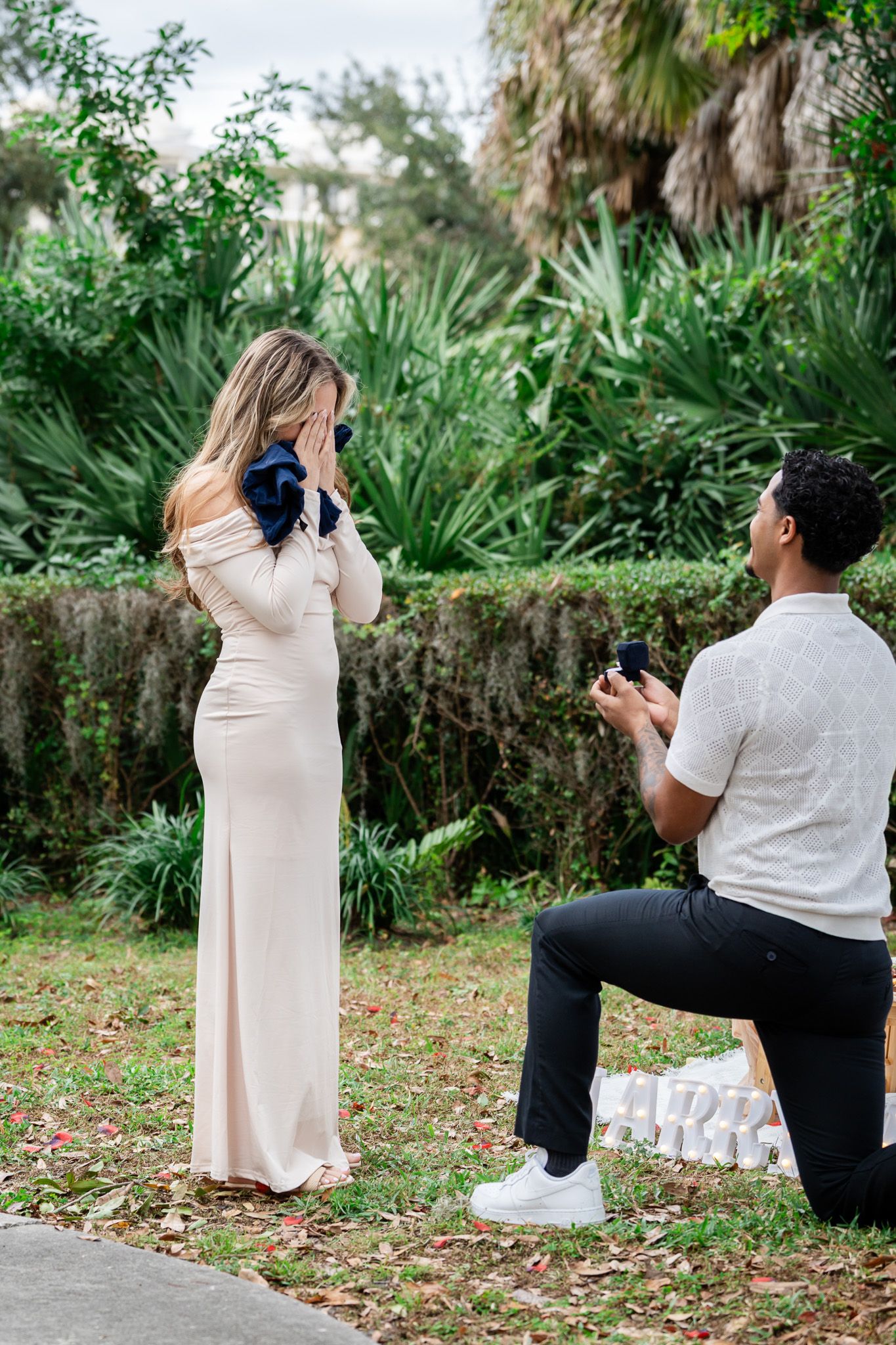 Emotional woman covers face as man proposes on one knee in tropical garden setting.