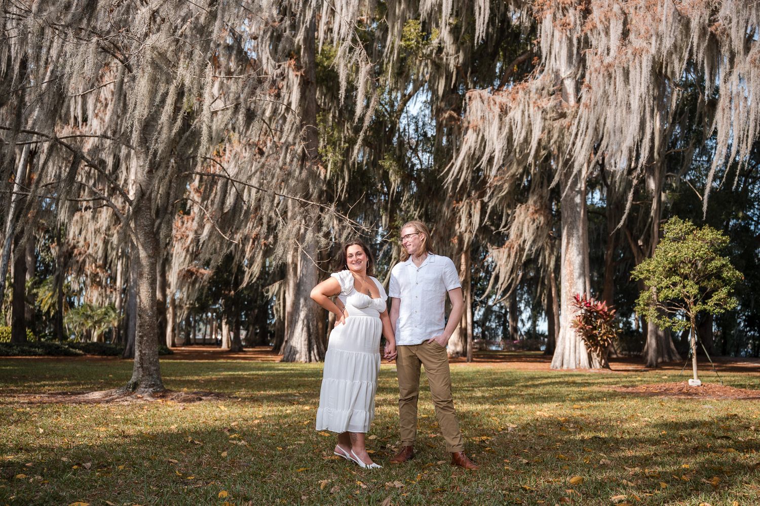 Smiling engaged couple stands together on green lawn beneath large Spanish moss-covered trees outdoors.