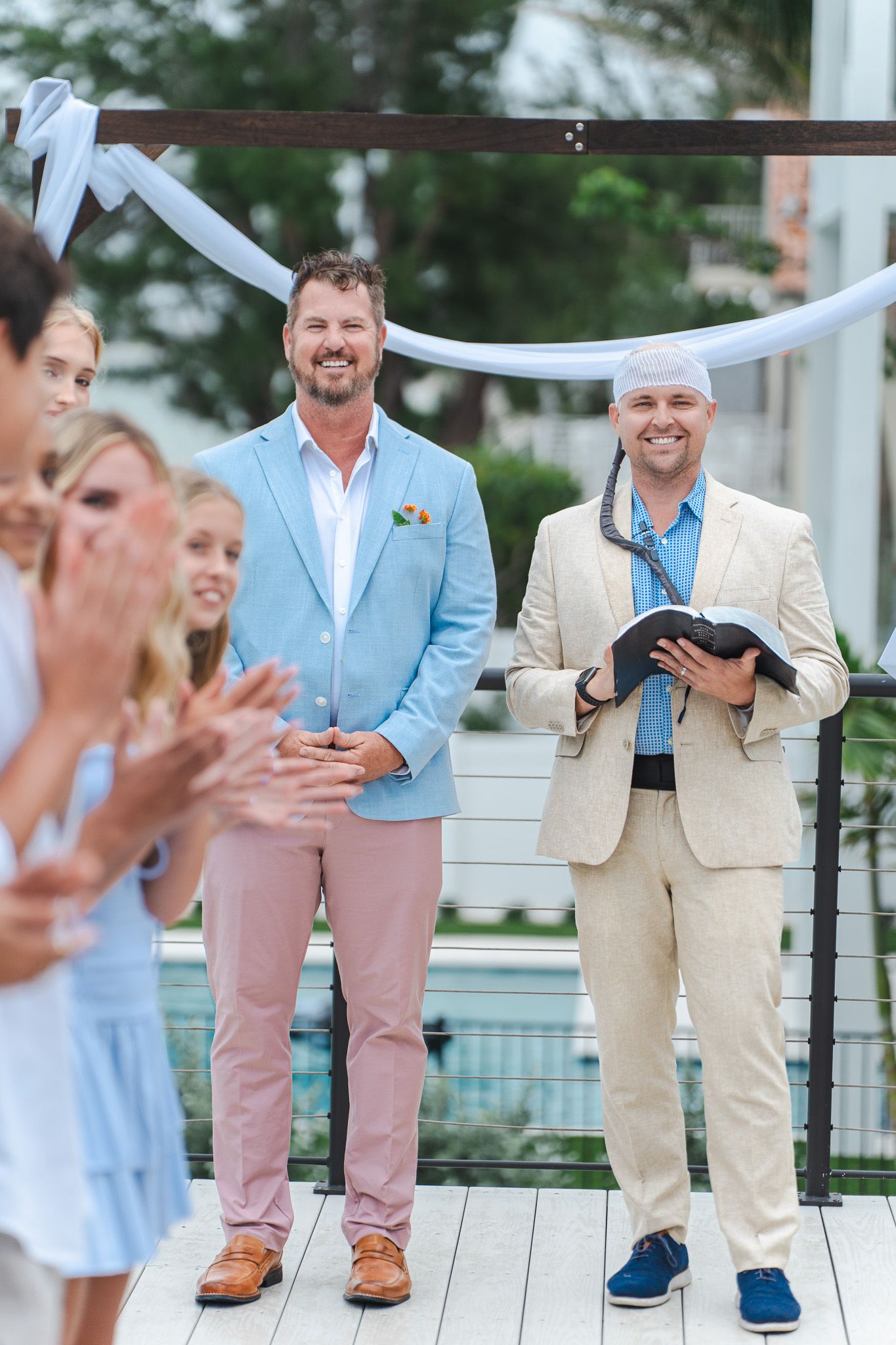 Groom in light blue blazer stands at wedding altar smiling as guests applaud during ceremony.