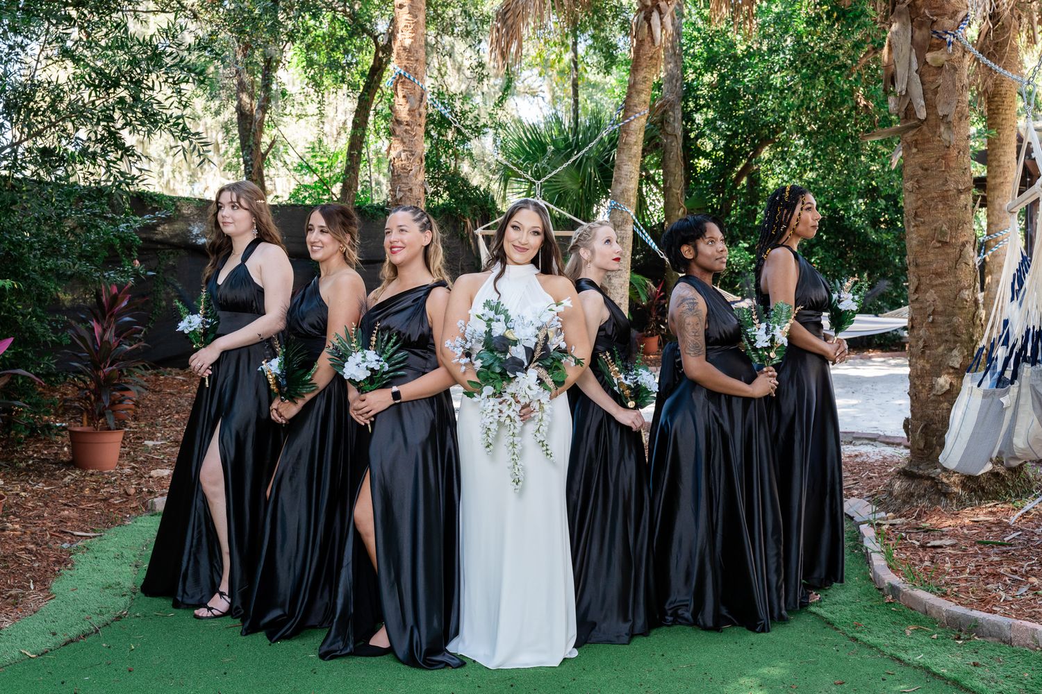 Bride in white gown stands with six bridesmaids in black satin dresses holding bouquets in garden.