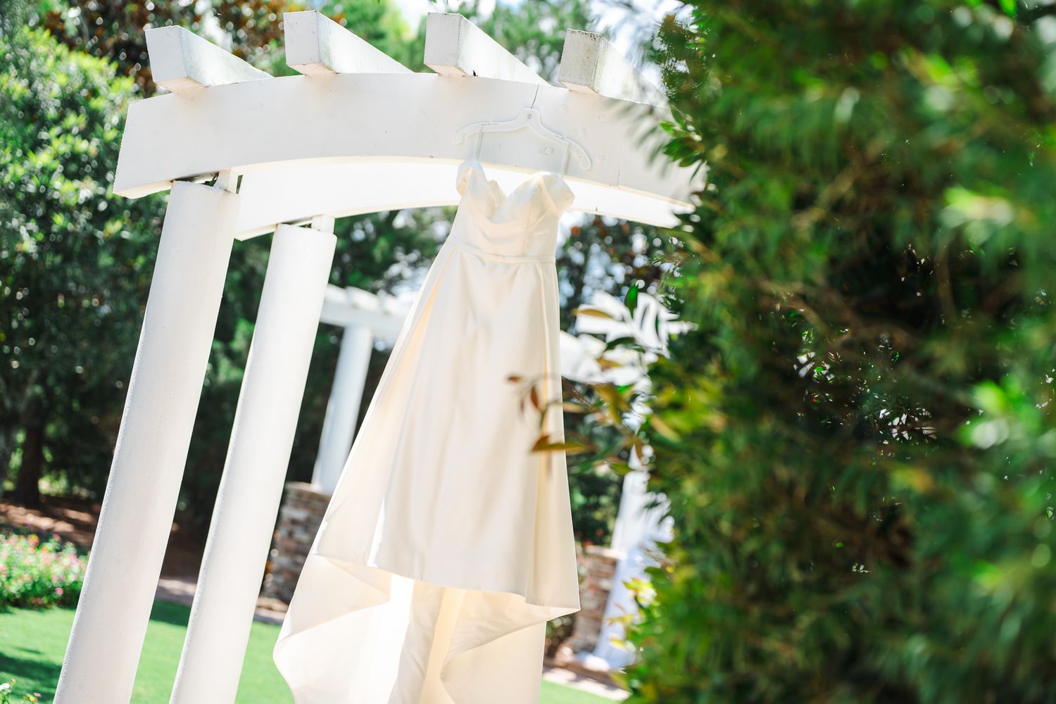 Elegant white wedding dress hanging from a white garden pergola surrounded by lush green trees.