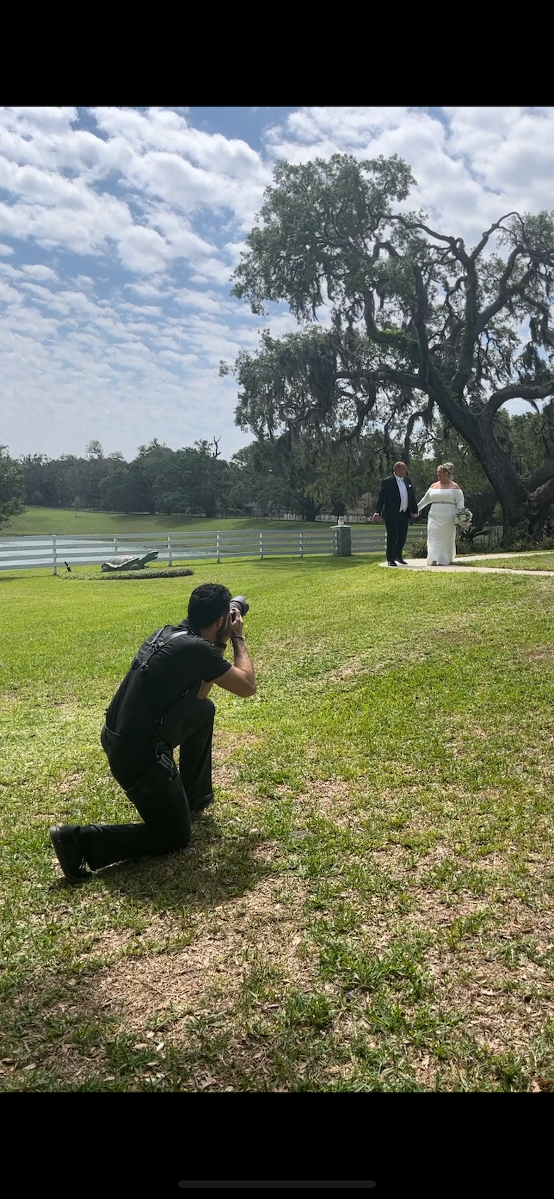 Wedding photographer kneeling on grass capturing bride and groom posing near a scenic lakeside oak tree.